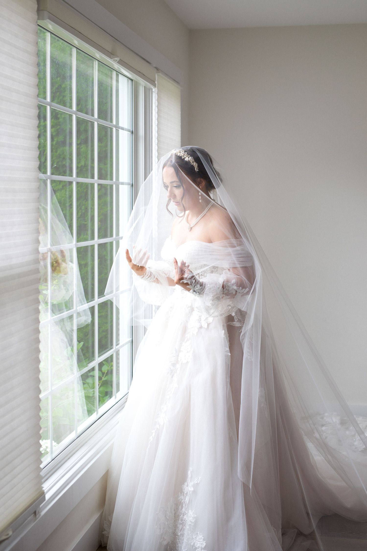 A flowing white wedding dress with delicate tulle veil captured in soft natural light through large windows.