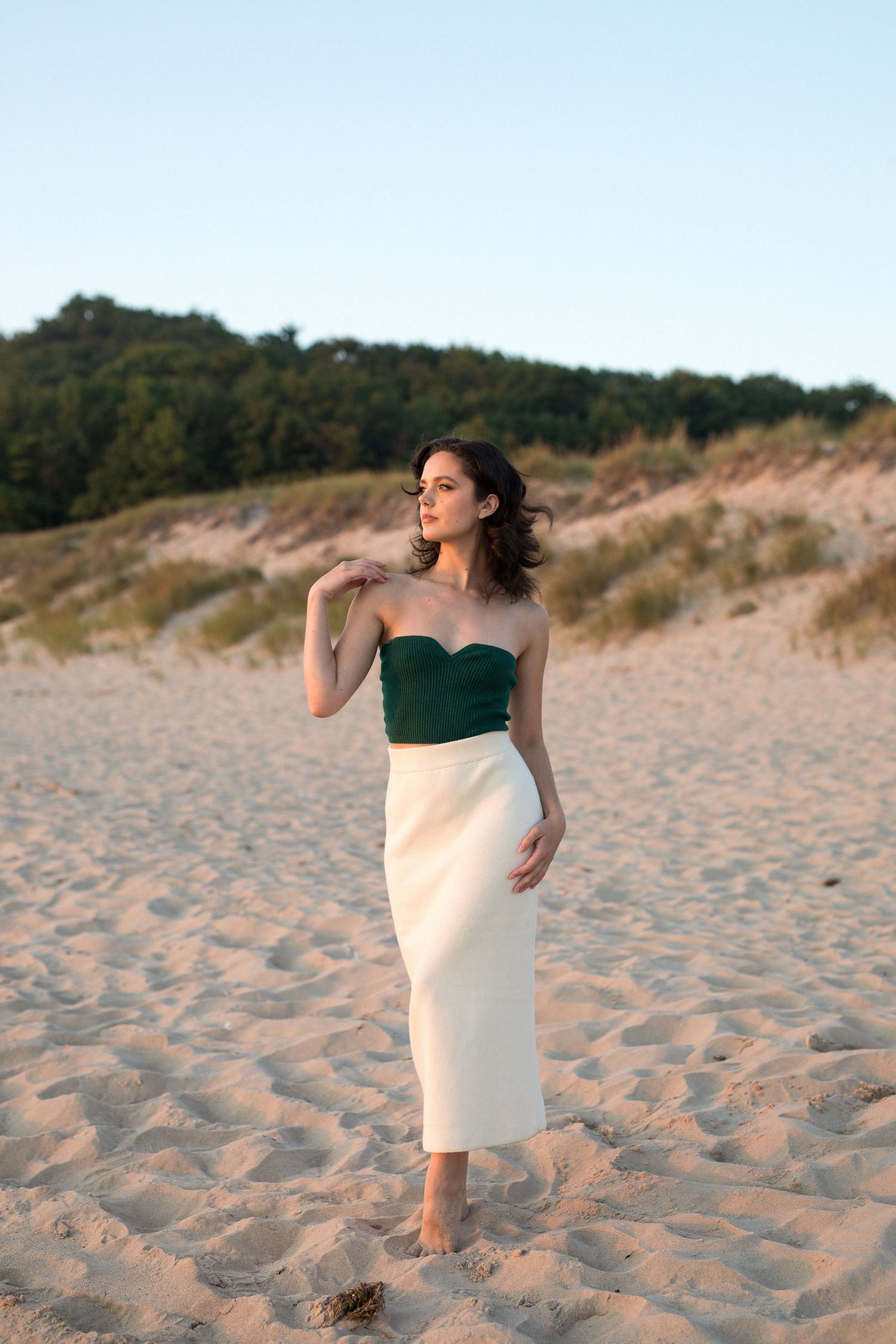 Woman in elegant two-tone dress walks along sandy beach with dunes and trees in background.