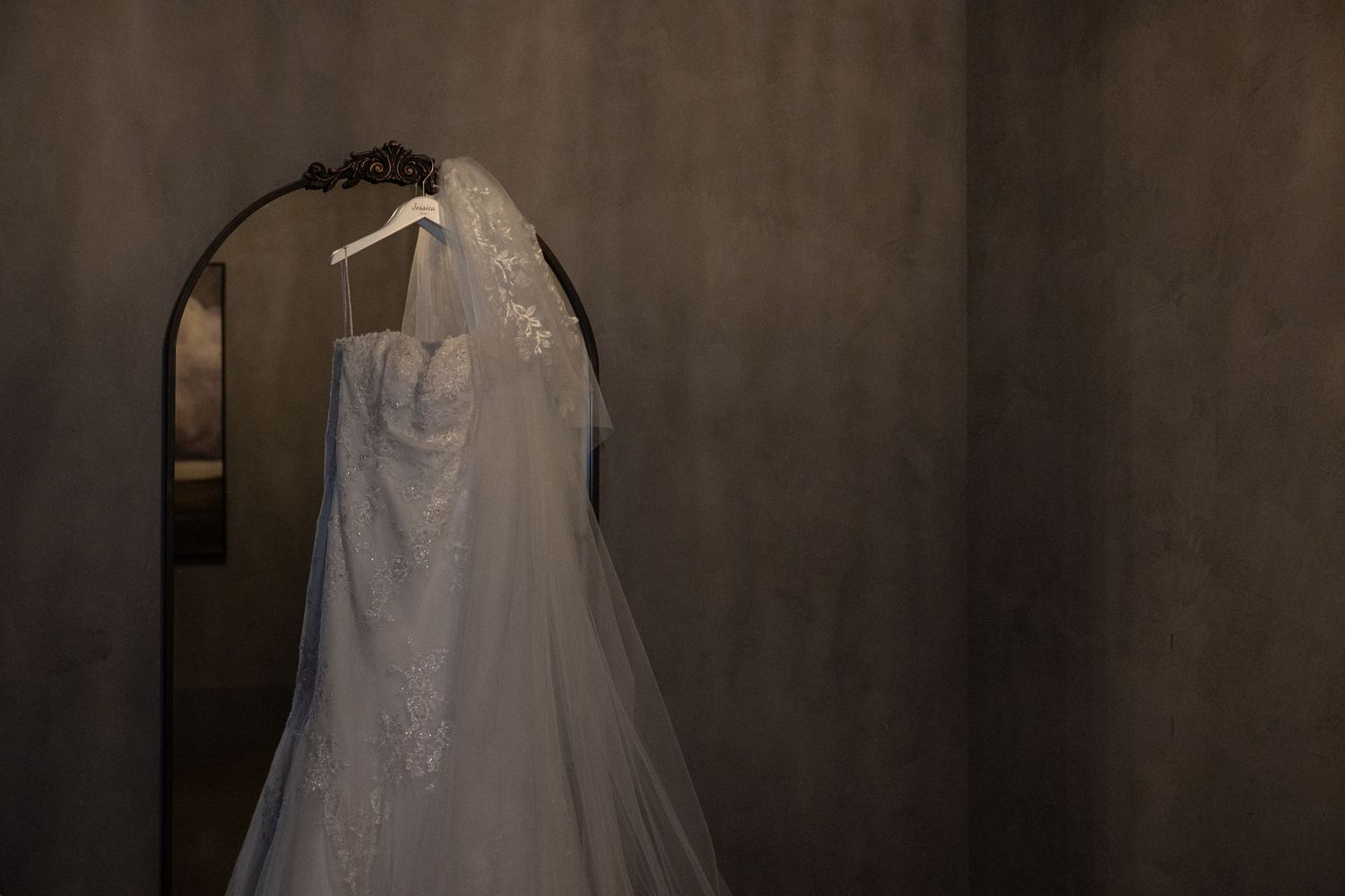 Elegant white wedding dress with lace veil hanging on a mirror in a dimly lit gray room.