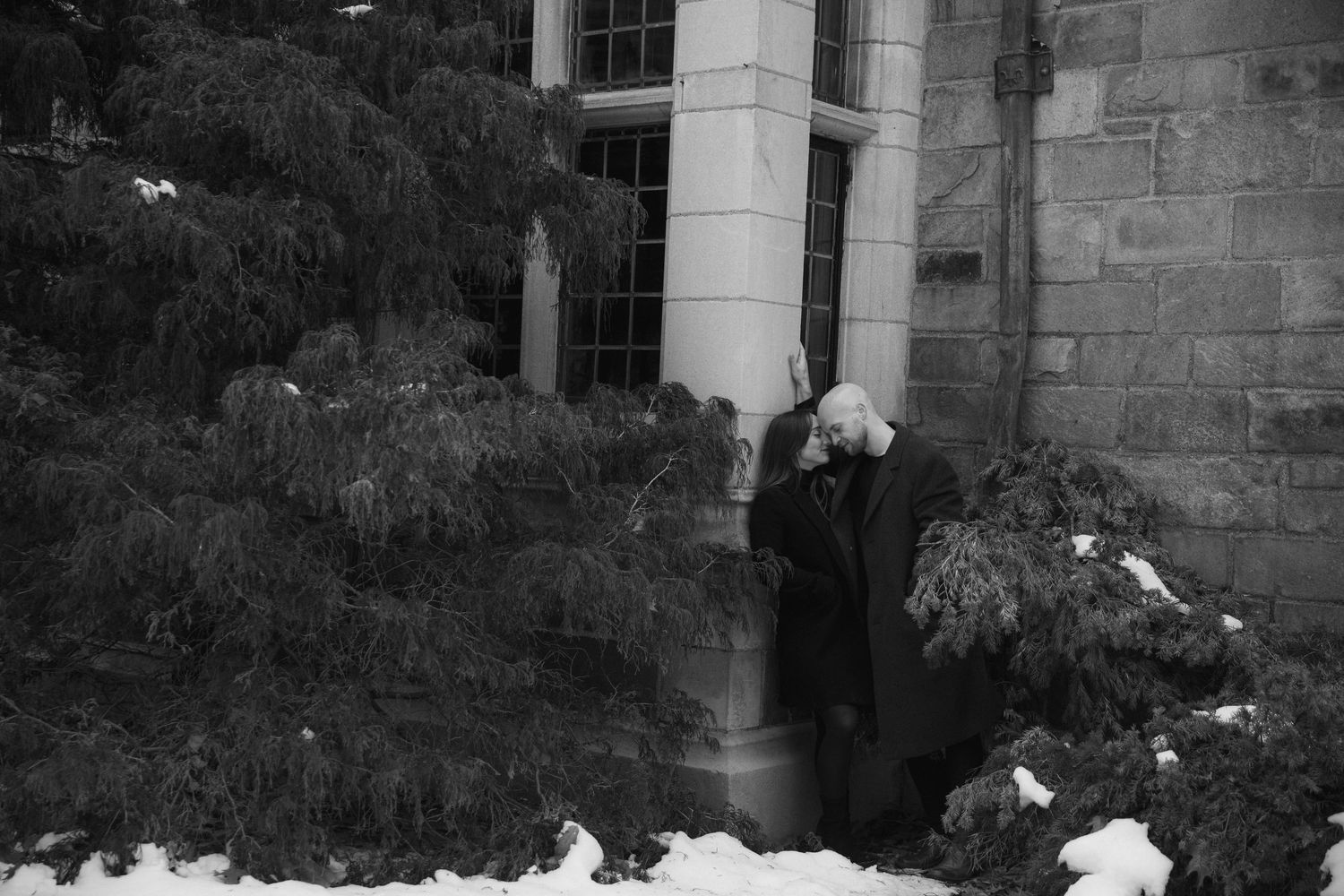 A figure in dark clothing stands in the doorway of a stone building surrounded by snow and foliage.