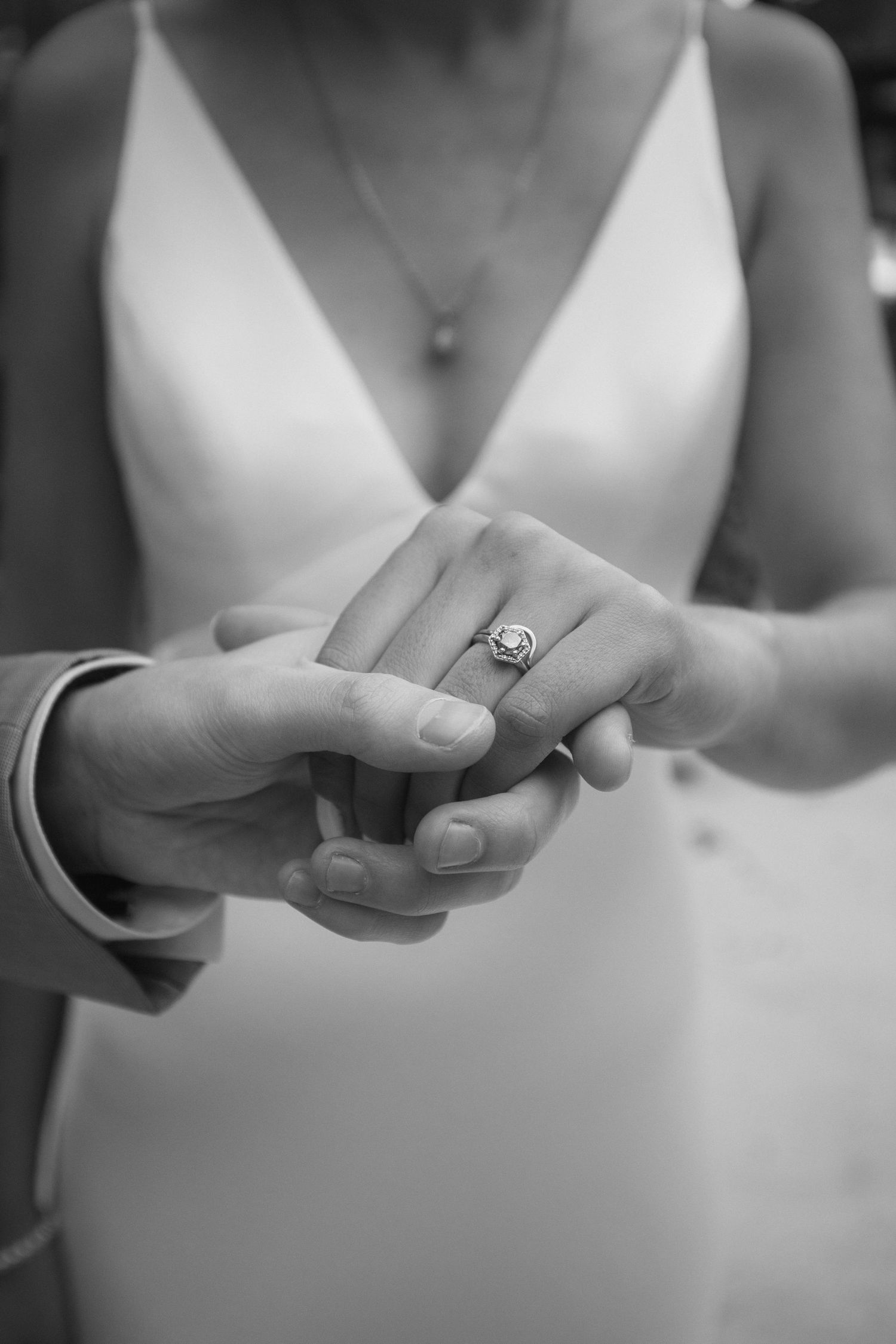 A black and white close-up of hands exchanging wedding rings during a ceremony, with a white dress visible in the background.