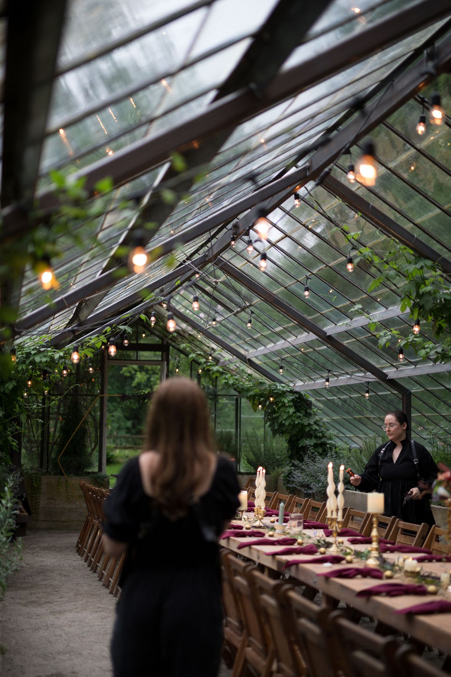 A rustic greenhouse wedding reception setup with string lights and long wooden tables decorated in burgundy accents.