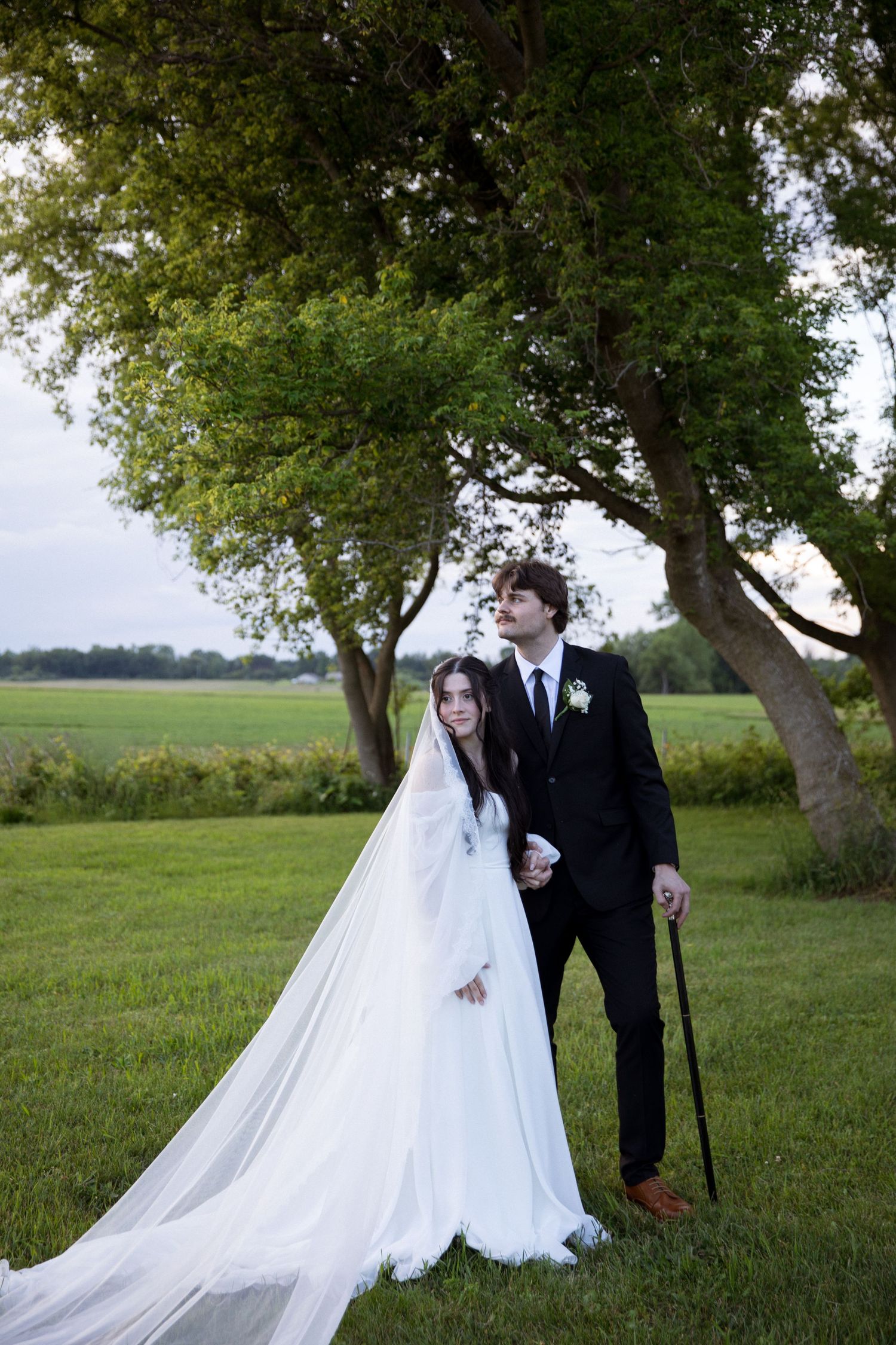 Bride in white gown with long veil and groom in black suit holding cane pose outdoors near trees on green lawn.