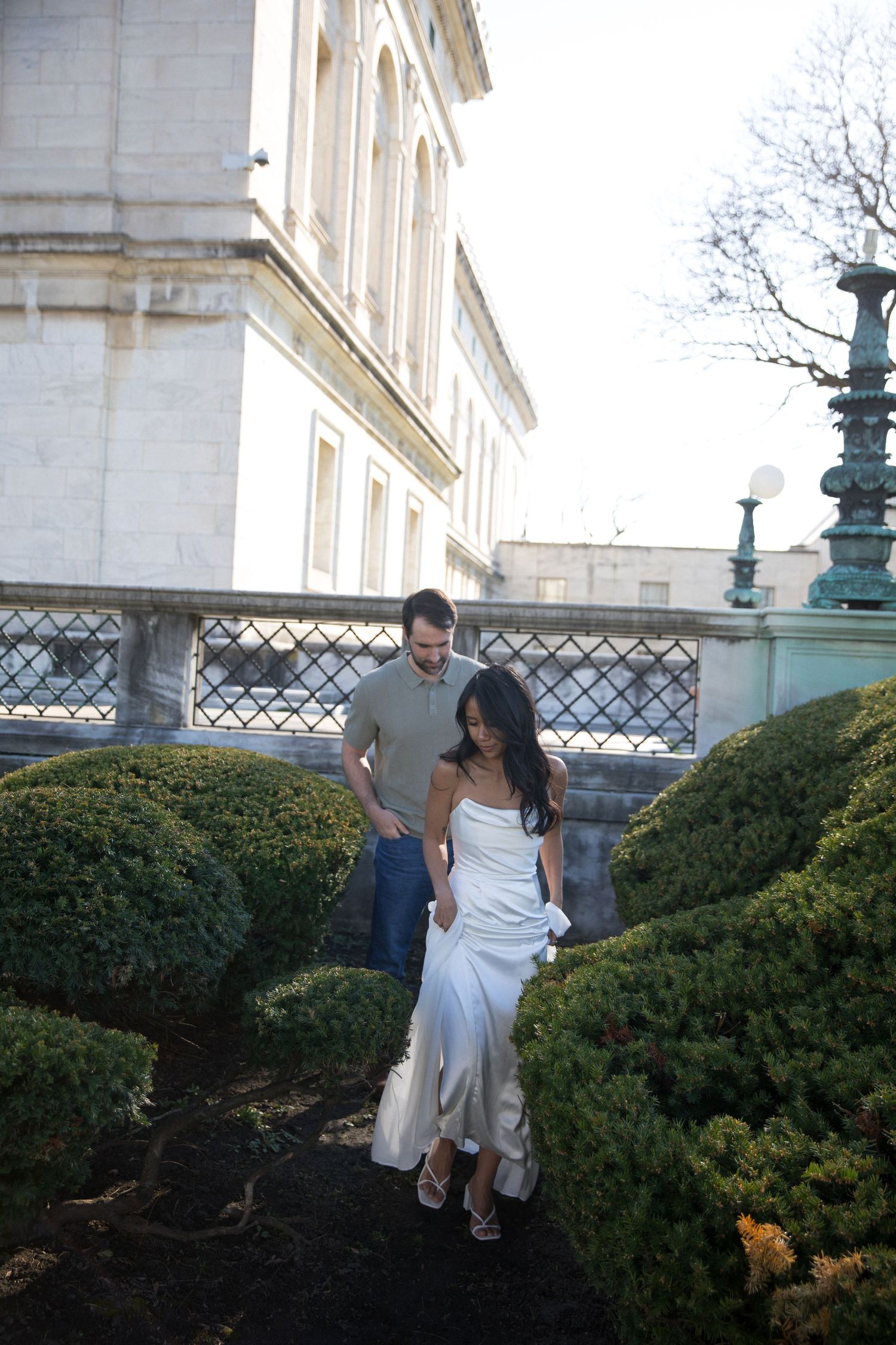 Couple walking through manicured garden hedges near elegant stone building in wedding attire.