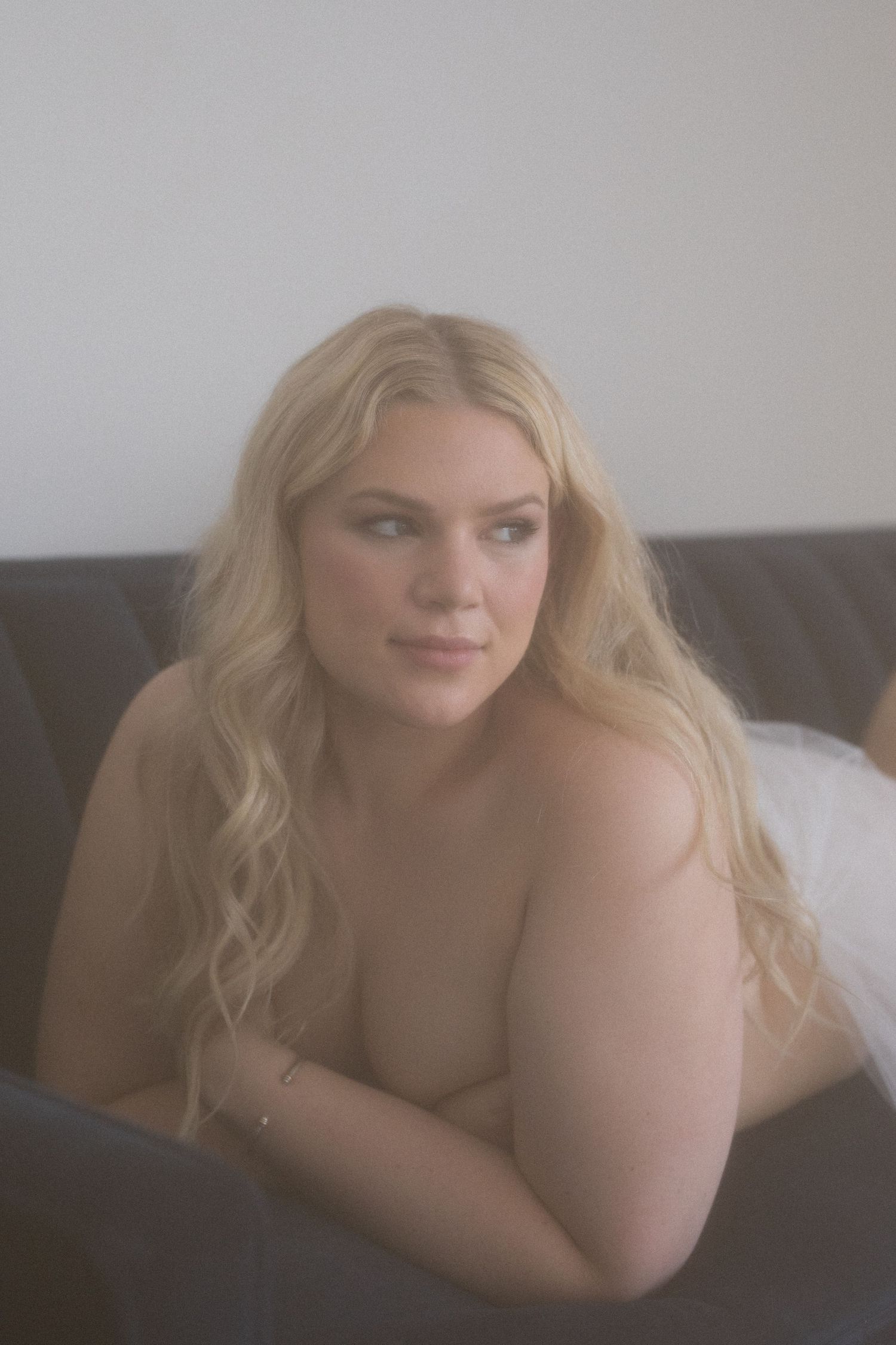 A young blonde woman with long wavy hair reclines on a dark sofa in a softly lit room.