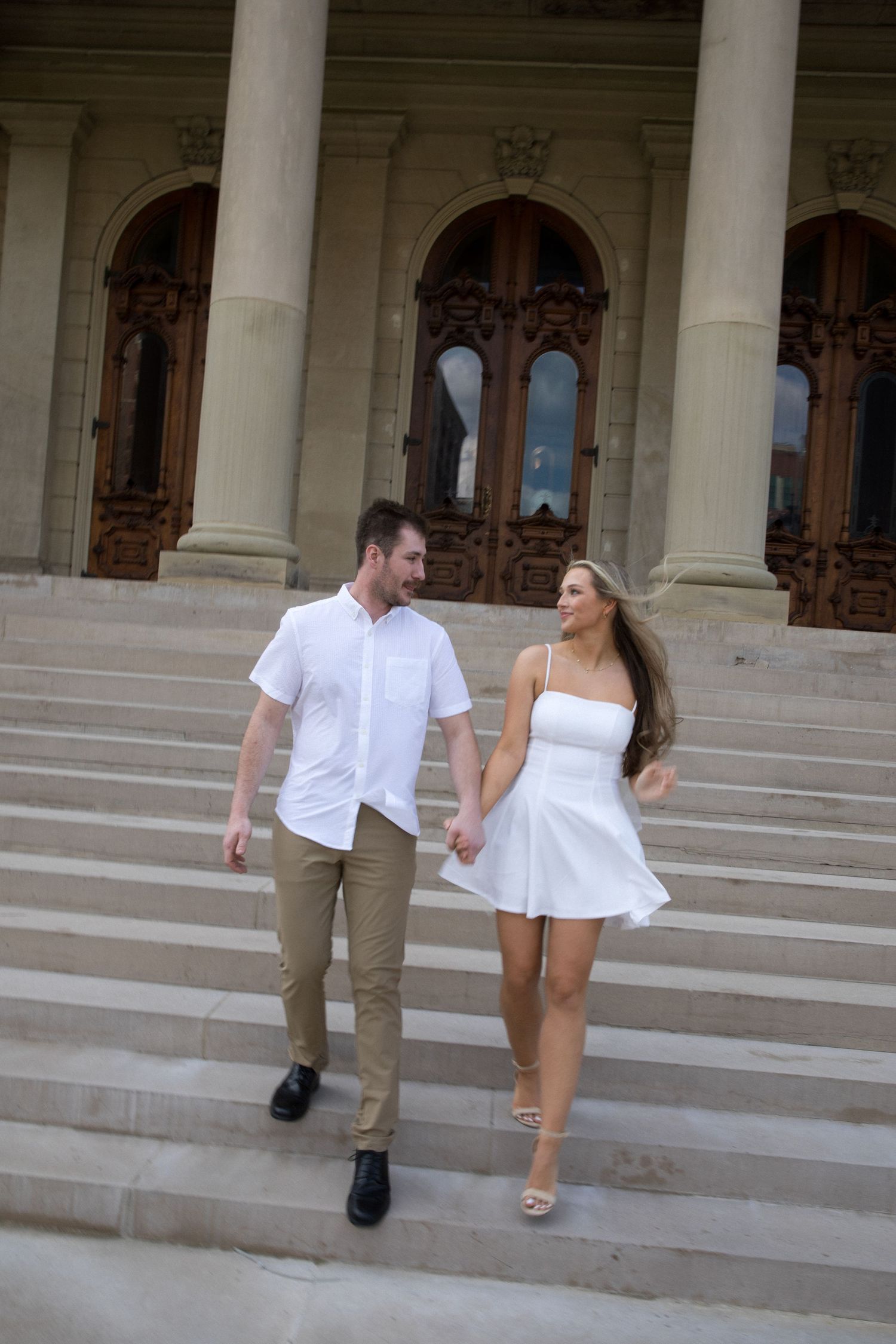 A smiling couple holding hands descending stone steps outside a grand classical building.