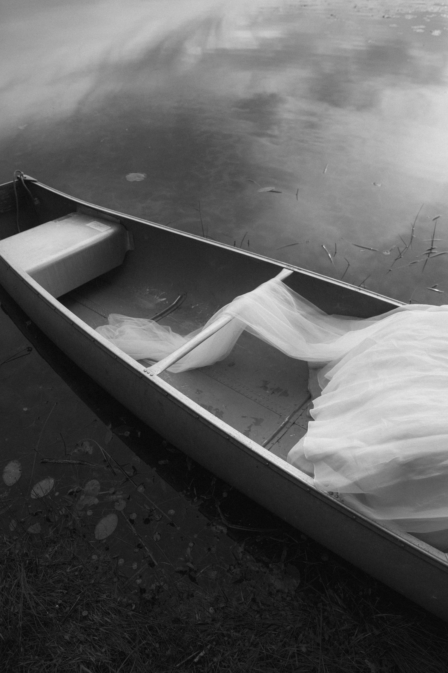 Black and white photo of a canoe at waters edge with white bridal gown fabric draped inside.