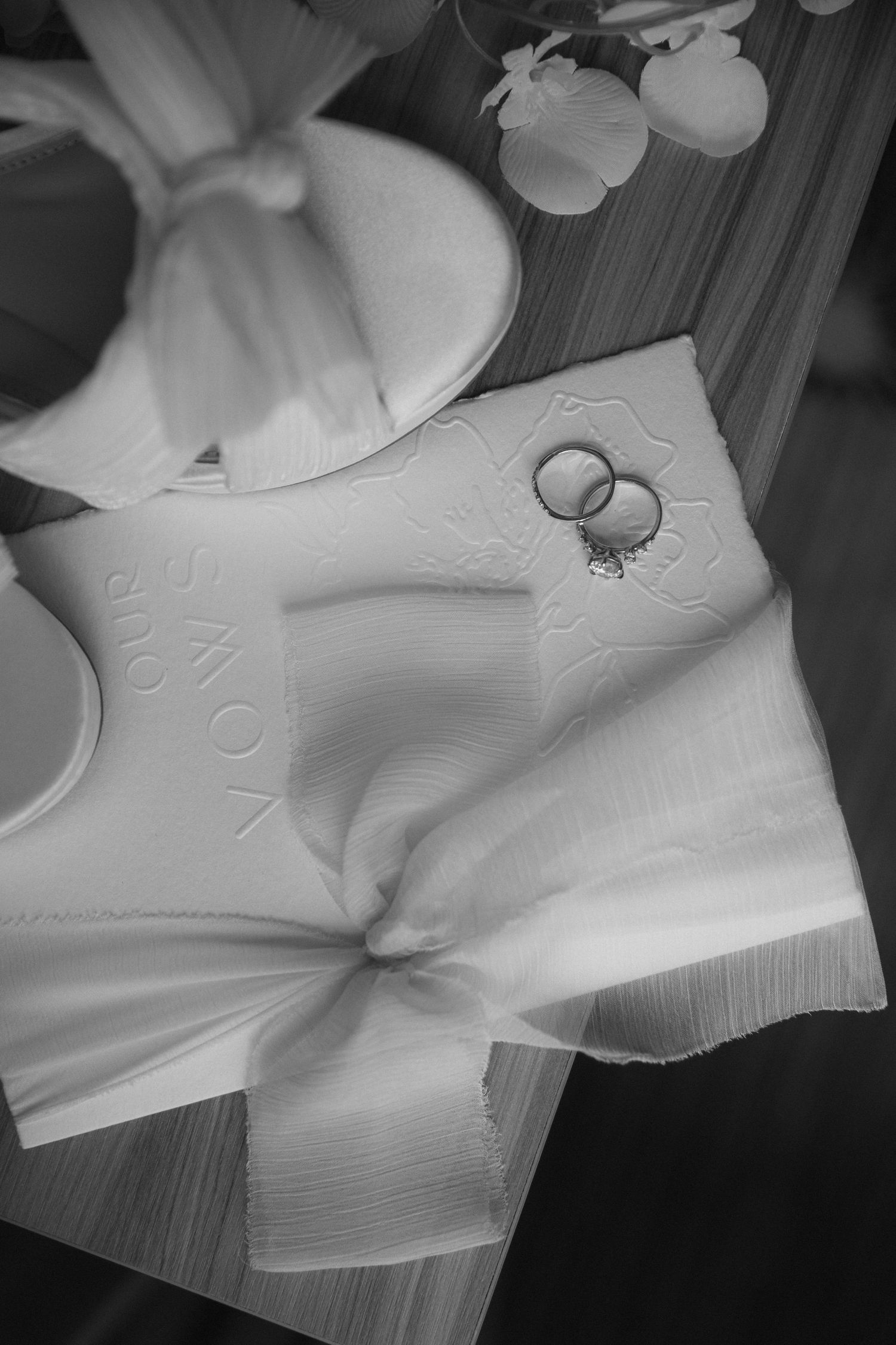 Wedding rings rest on a white napkin or fabric with delicate floral decor in black and white photography.