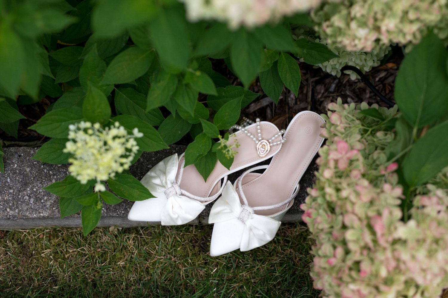A pair of elegant white bridal shoes with delicate bows rests among green foliage and pink flowers.