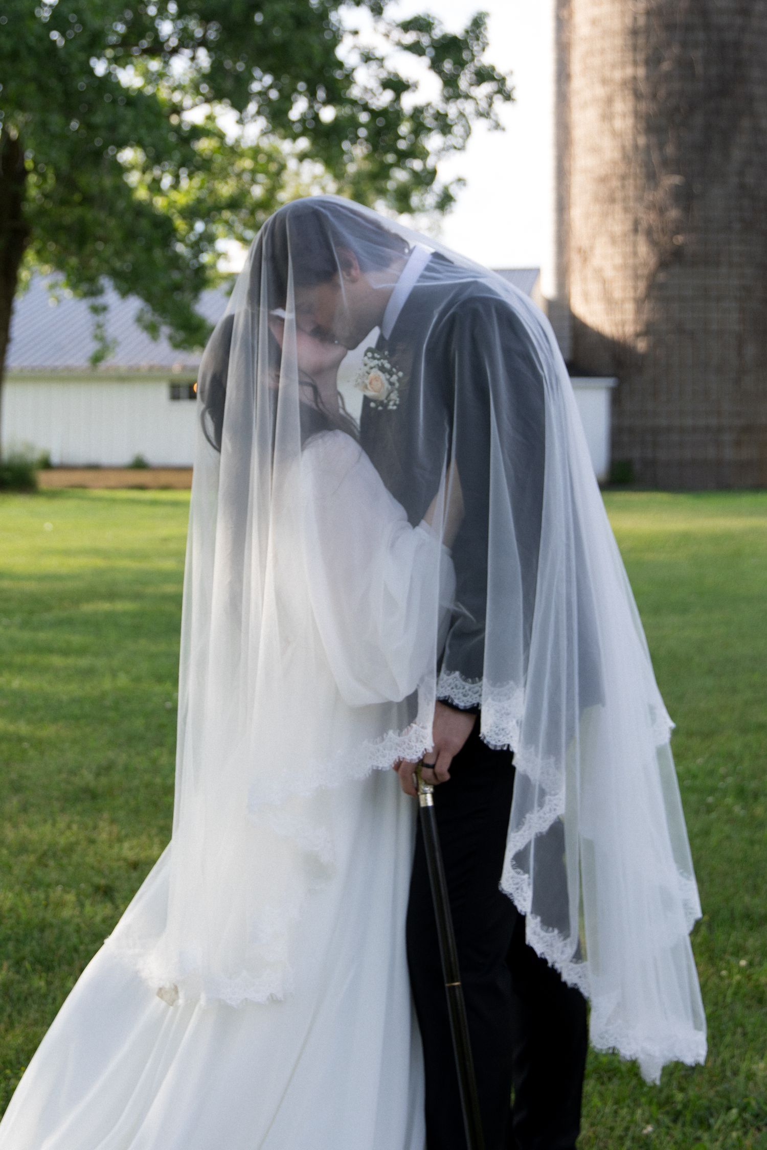 Bride and groom sharing a kiss under a white wedding veil outdoors near a barn on their wedding day.