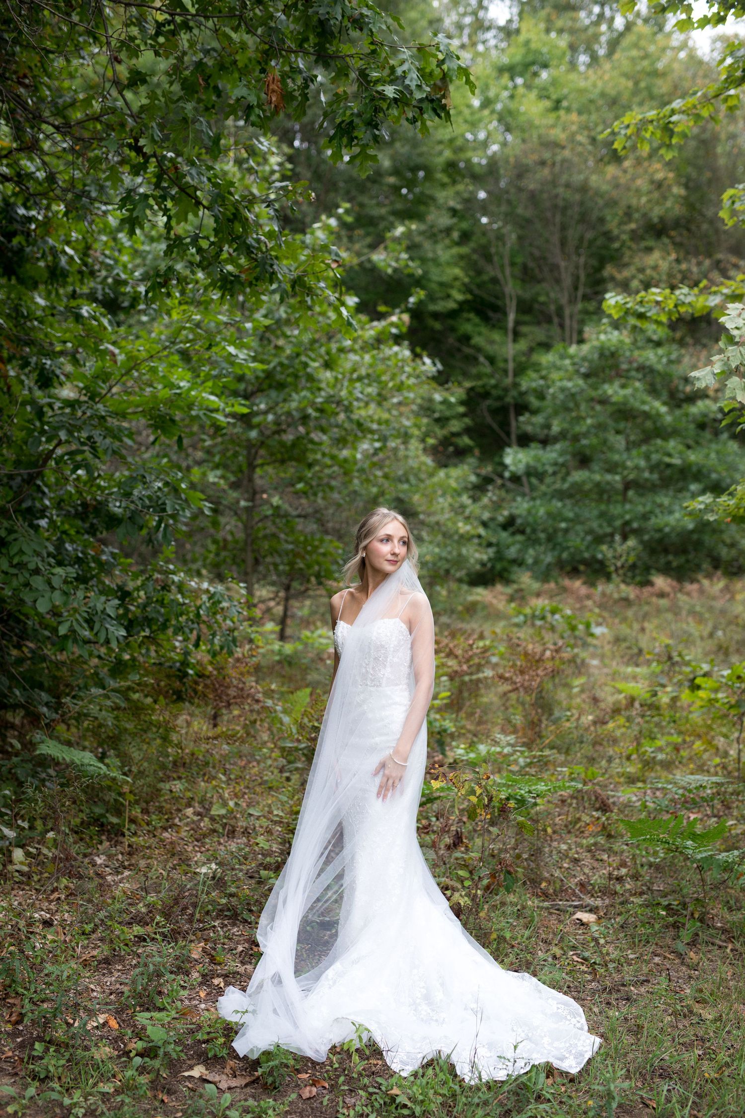 A flowing white lace wedding dress twirls gracefully in a wooded outdoor setting during a bridal photoshoot.