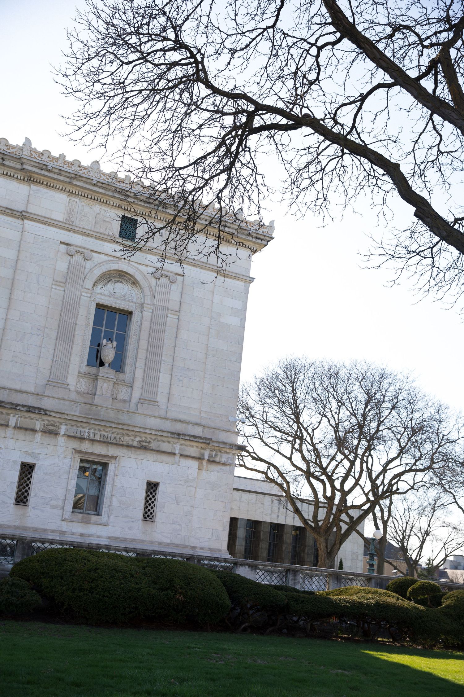 Historic white marble neoclassical building with ornate architecture surrounded by bare winter trees and manicured green hedges.