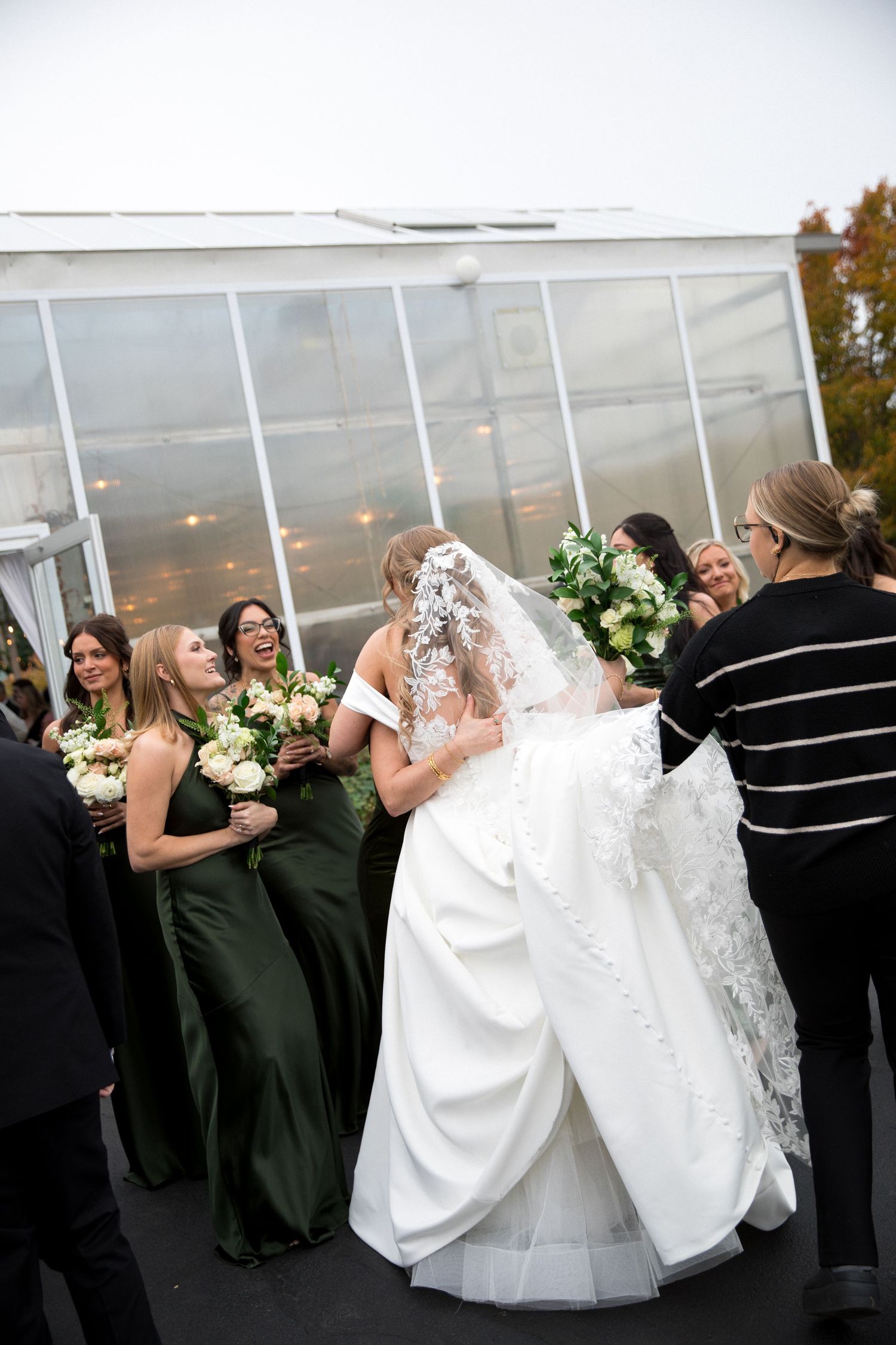 Wedding party in dark dresses and white gowns celebrating together outdoors near a greenhouse.