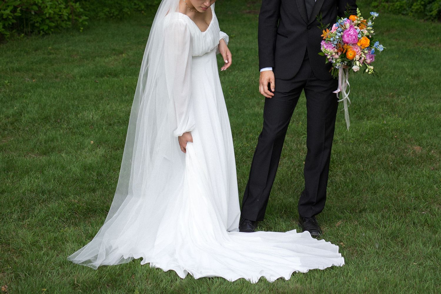Wedding couple stands in a lush green garden with the bride in a flowing white gown and groom in a dark suit.