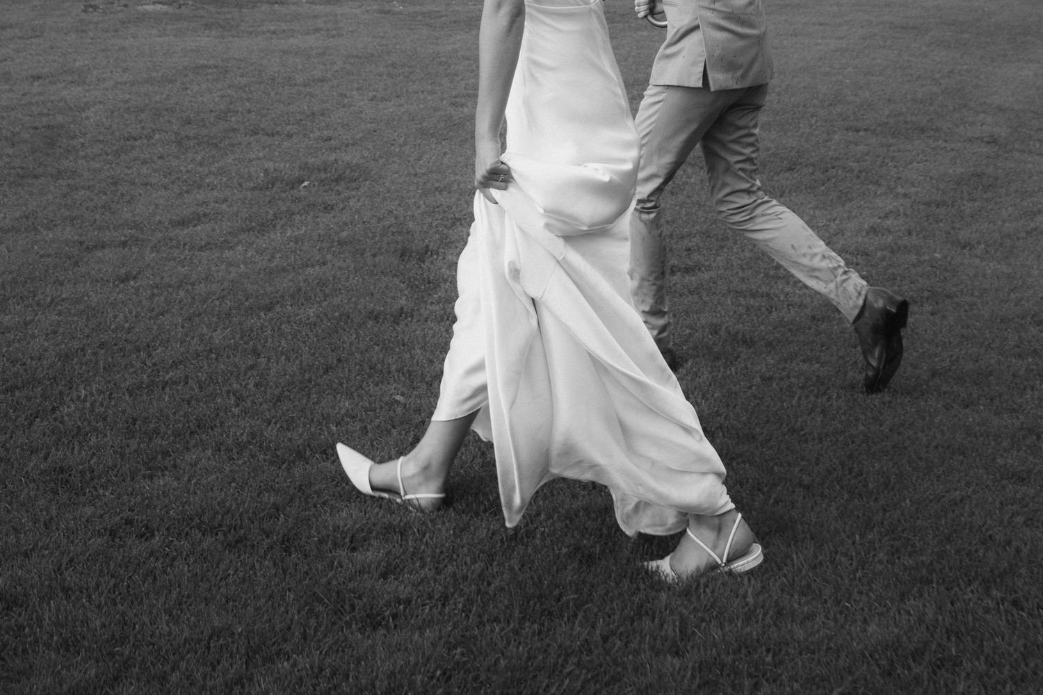 Black and white photo of bride and groom walking on grass, showing dress movement and shoes.