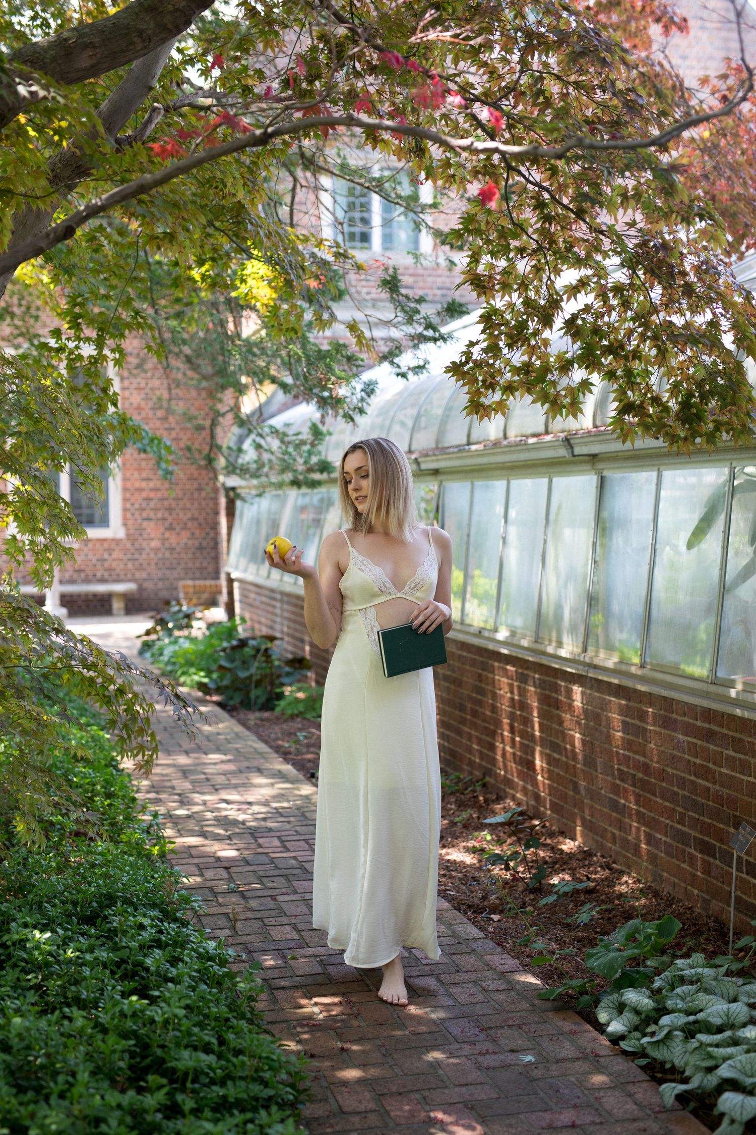 A person in a light dress walks along a brick pathway bordered by green foliage and trees with autumn colors.