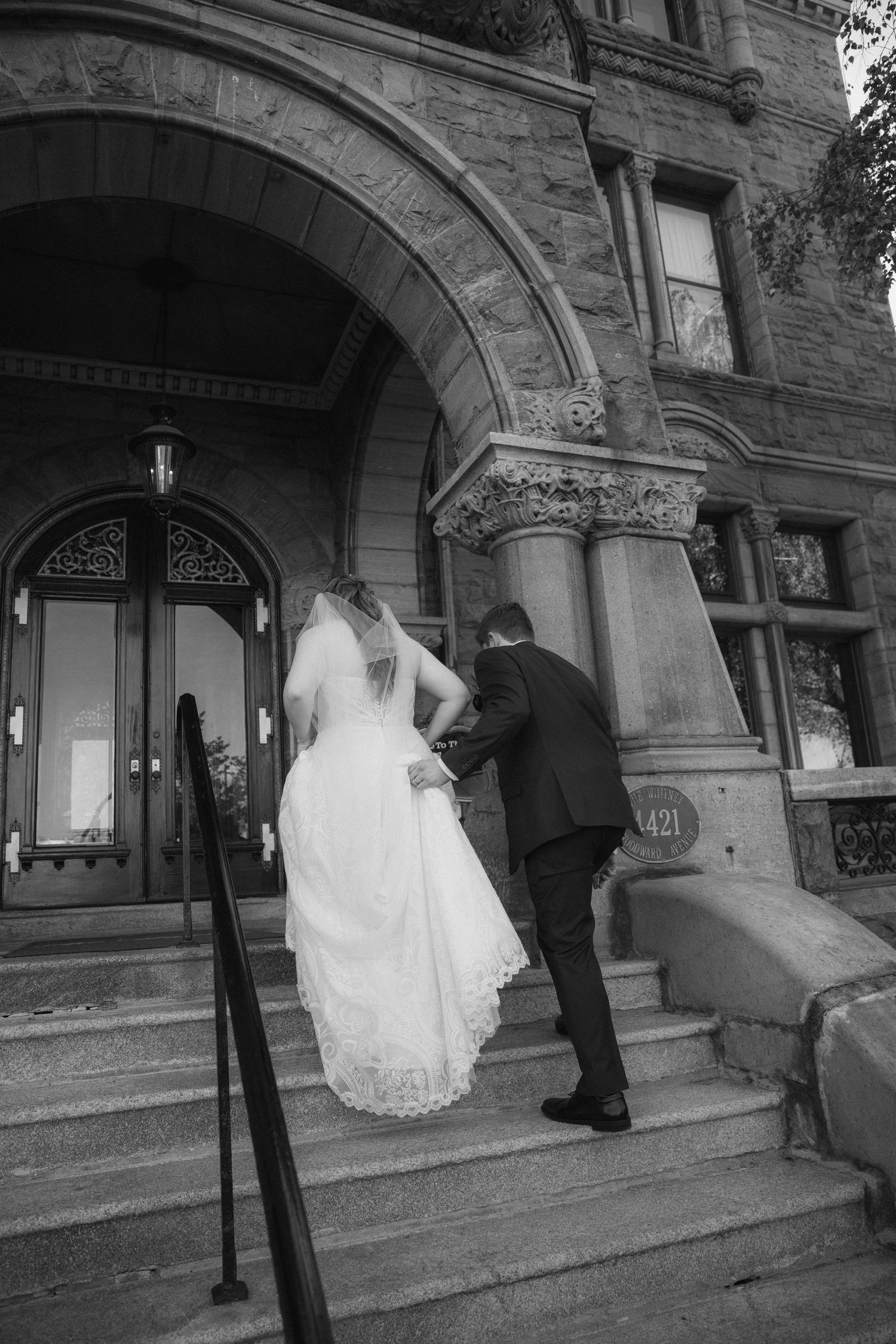 A couple in wedding attire climbing stone steps at the entrance of a historic building with ornate architectural details.