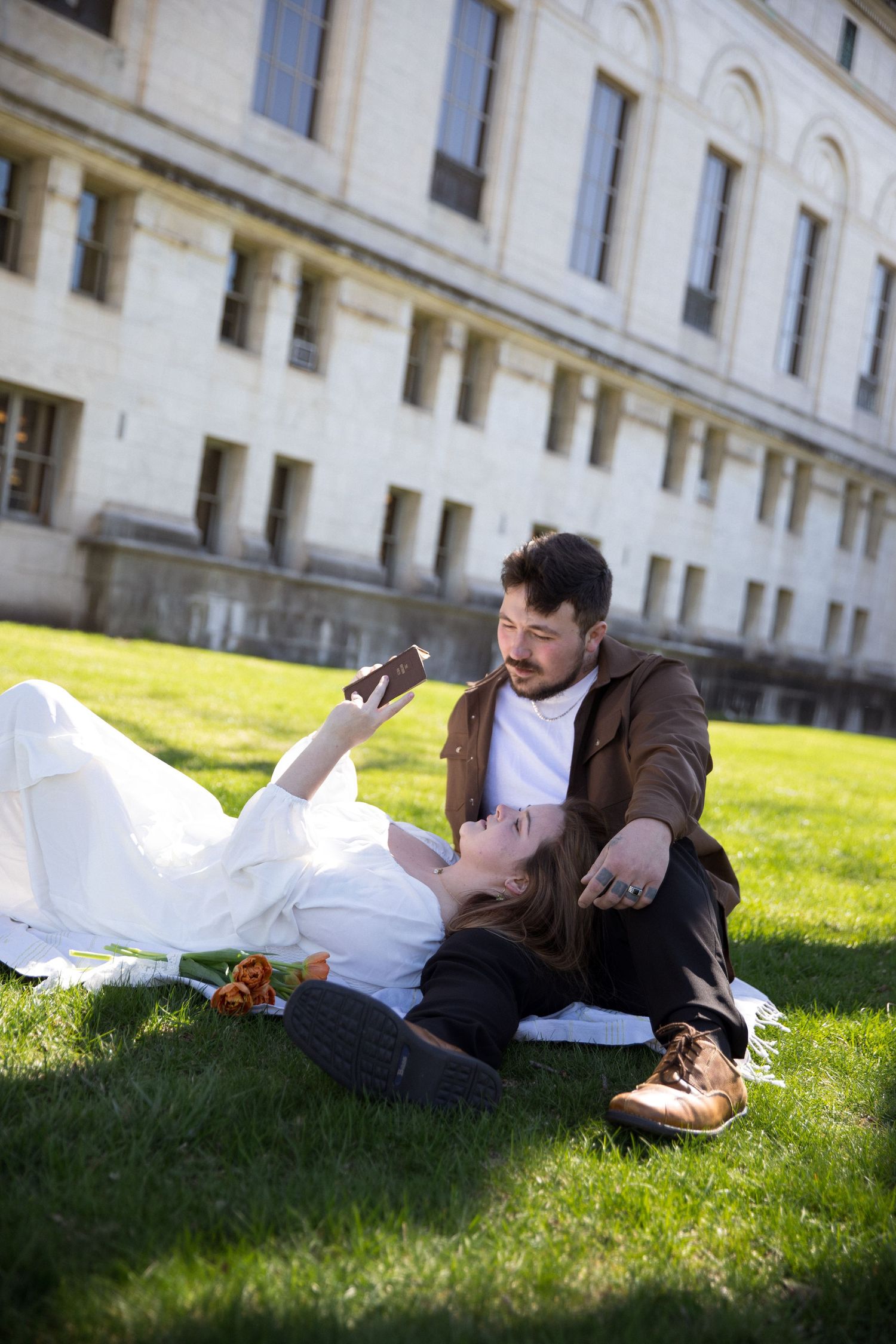 Couple relaxing on green grass near historic building, woman lying down showing phone to seated man.