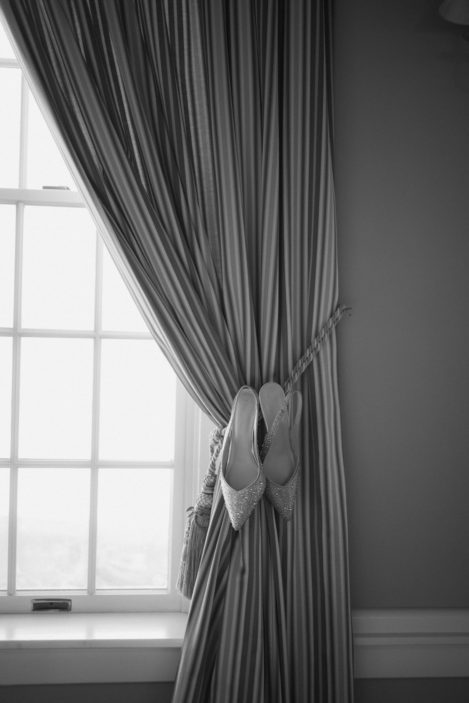 A pair of elegant white bridal shoes hangs decoratively on gray curtains by a bright window.