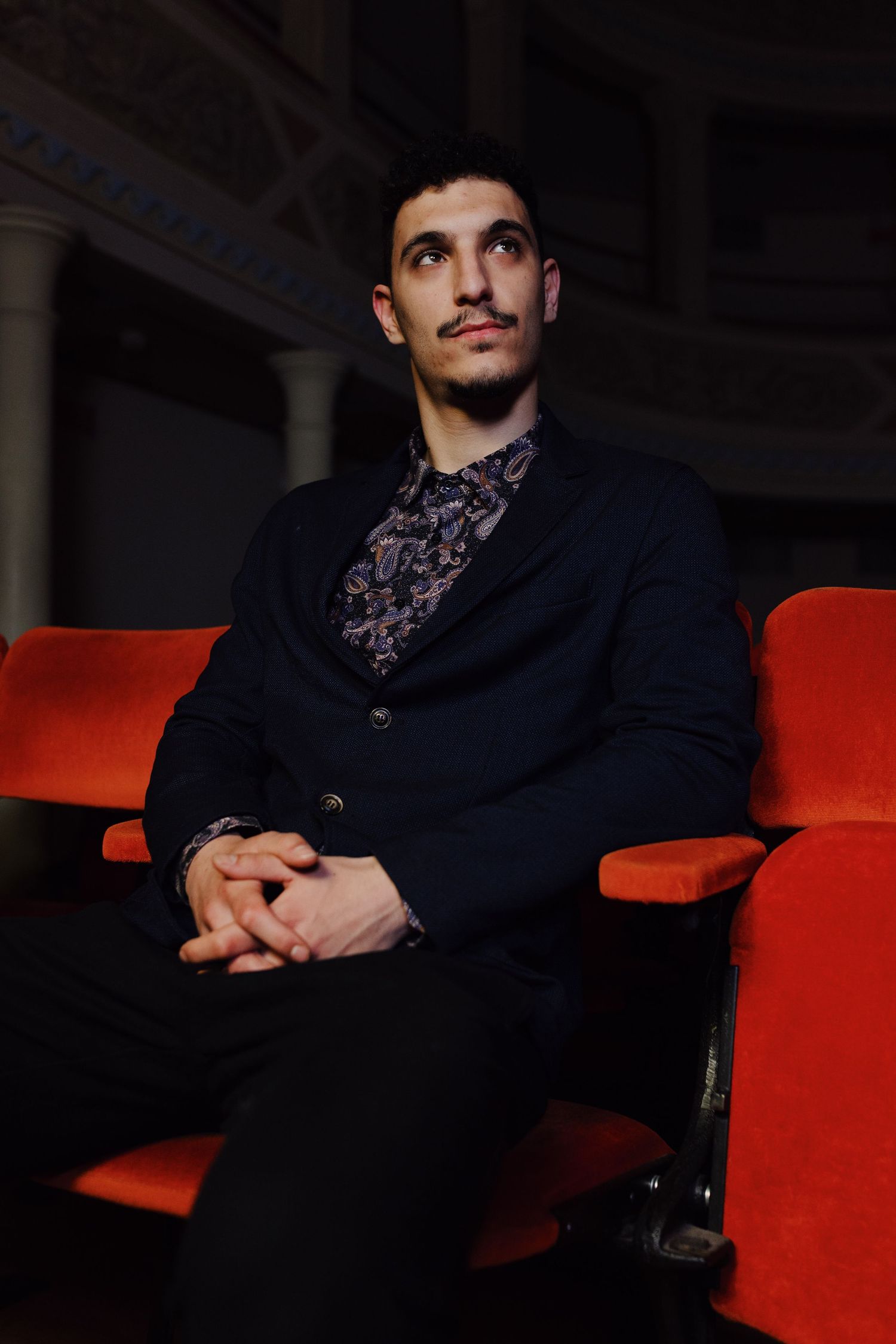 Un ragazzo vestito elegante è seduto su una poltrona rossa di teatro ed aspetta di essere chiamato per un provino.
