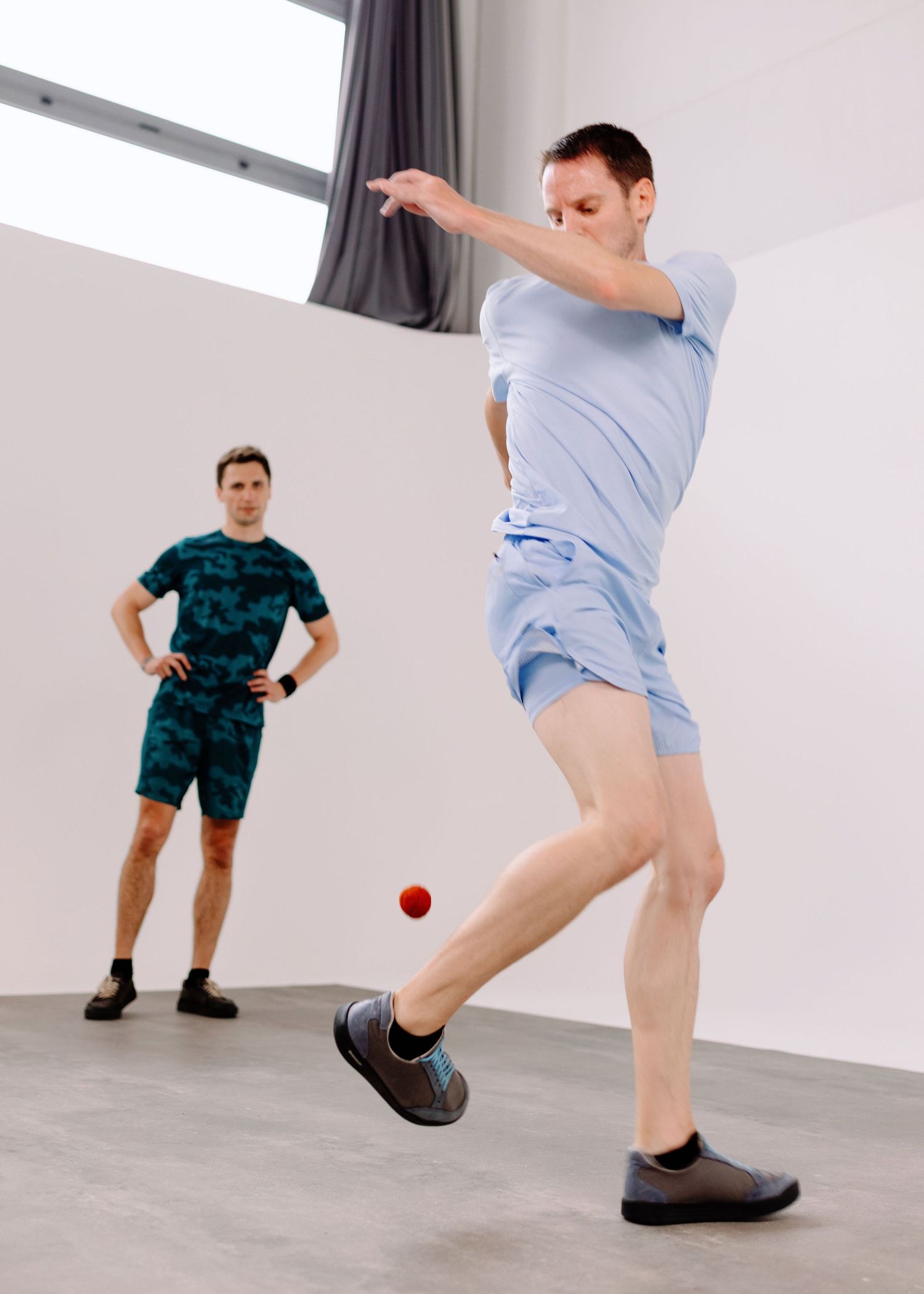 Due ragazzi vestiti di azzurro giocano allo sport footbag in una sala posa con un limbo bianco ed un pavimento grigio.
