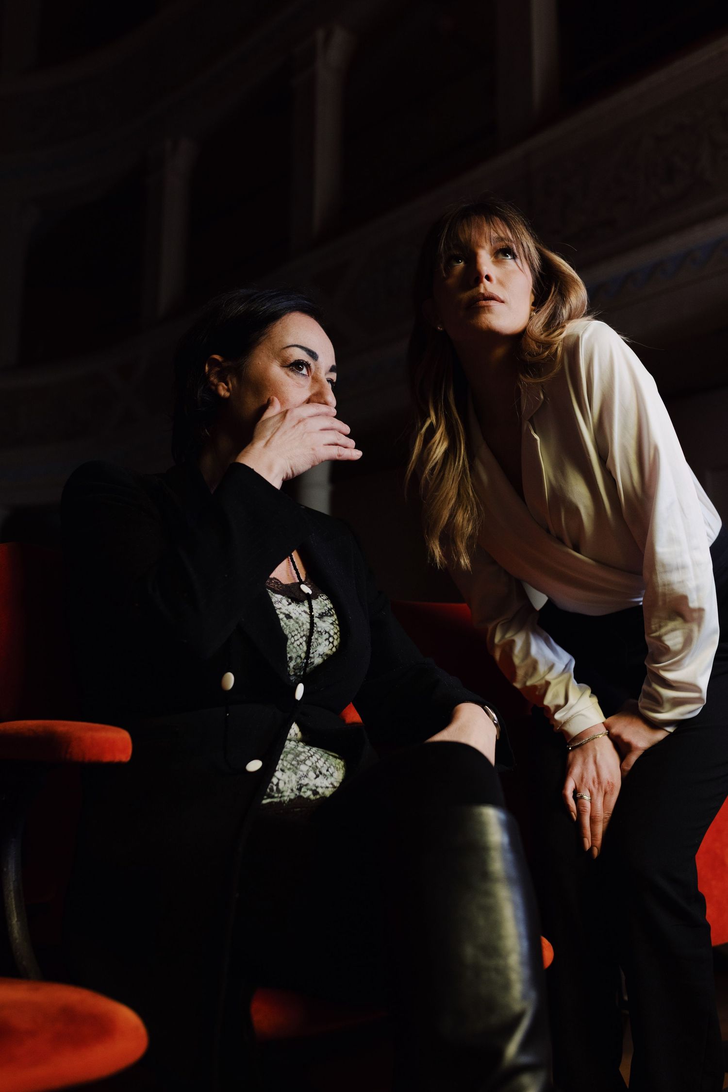 Due donne si sussurrano ciò che stanno vedendo parlandosi nelle orecchie in una stanza di teatro buia.