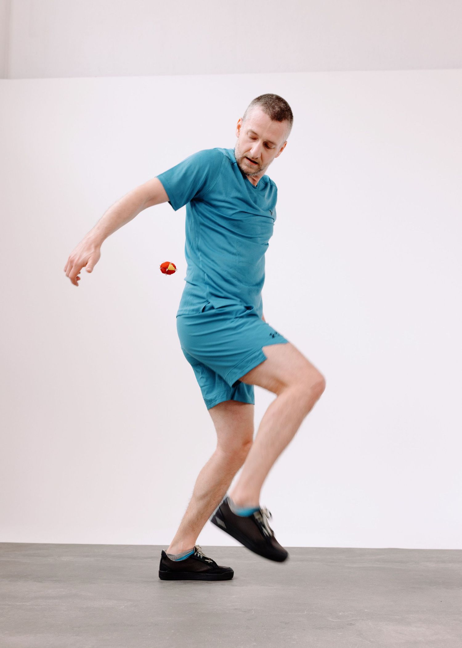 Un ragazzo con i capelli chiari vestito di verde acqua gioca allo sport footbag in una sala posa con un limbo bianco ed un pavimento grigio.