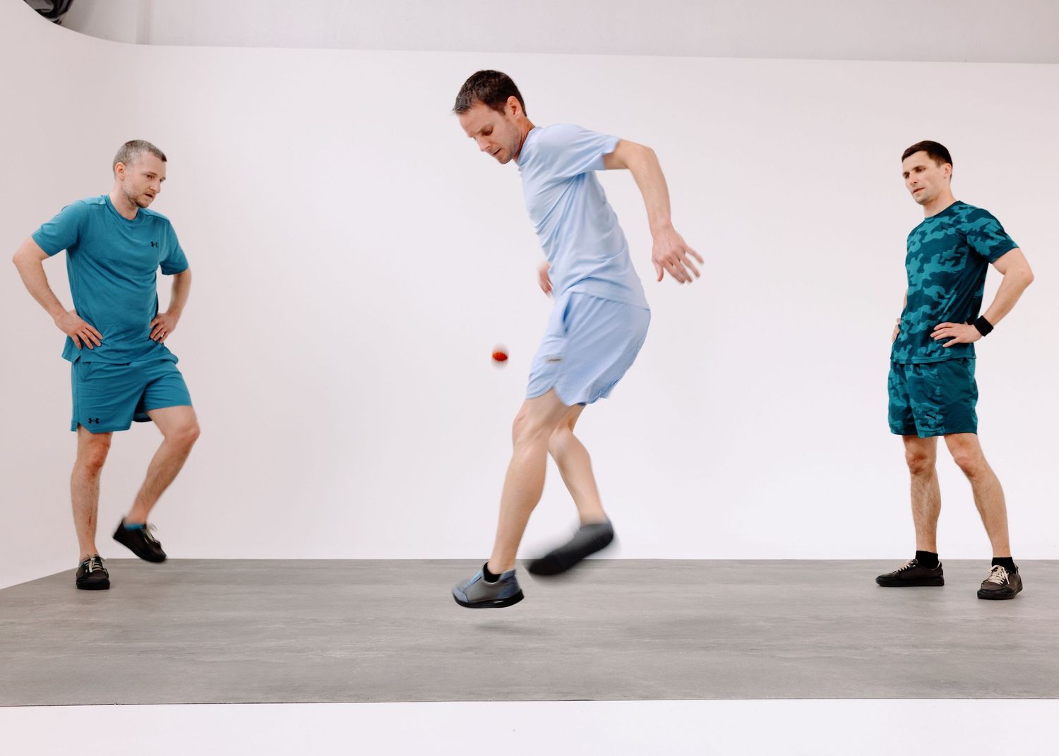 Tre ragazzi vestiti di azzurro giocano allo sport footbag in una sala posa con un limbo bianco ed un pavimento grigio.