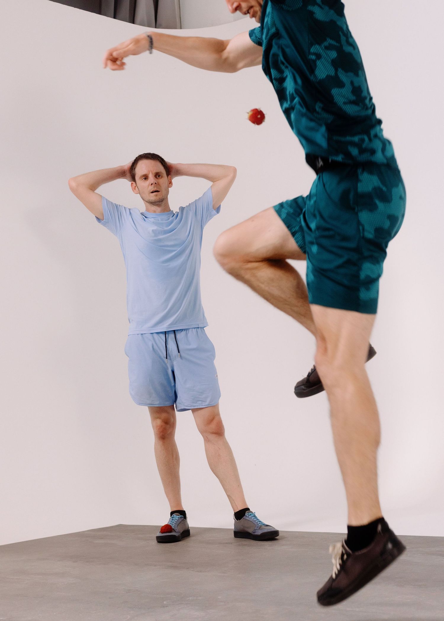 Due ragazzi vestiti di azzurro giocano allo sport footbag in una sala posa con un limbo bianco ed un pavimento grigio.