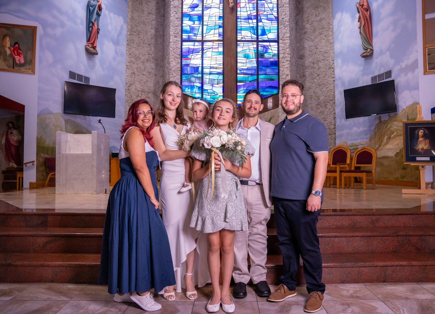 Bride, groom, and bridal party pose in church at alter as the flower girl holds the bride's bouquet.