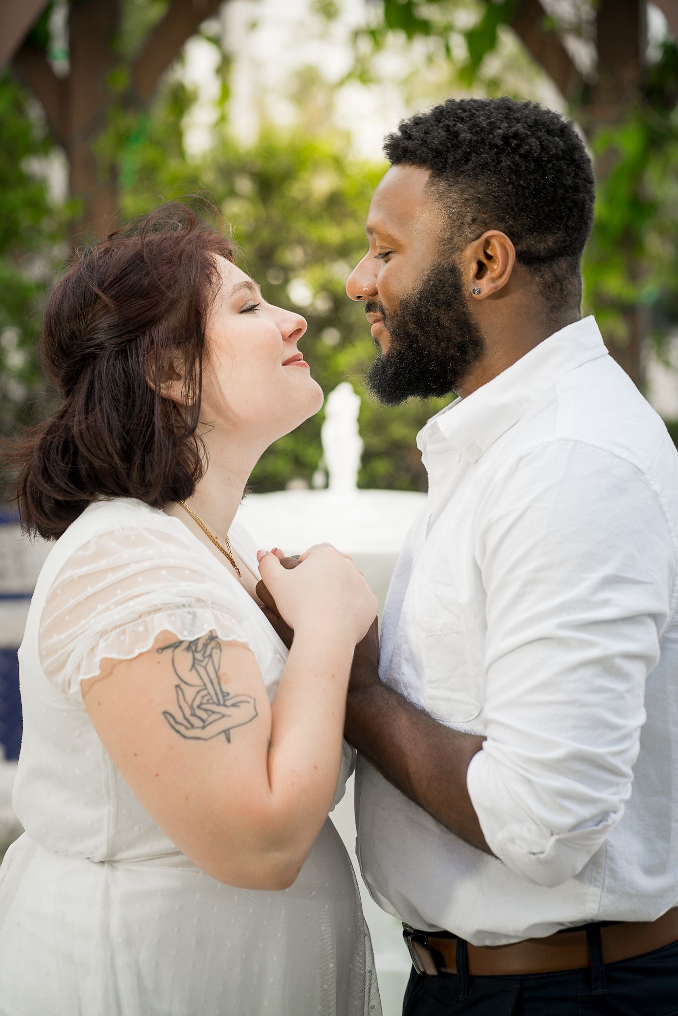 Couple gazes into each other's eyes while holding hands. Greenery and trees out of focus in the background.