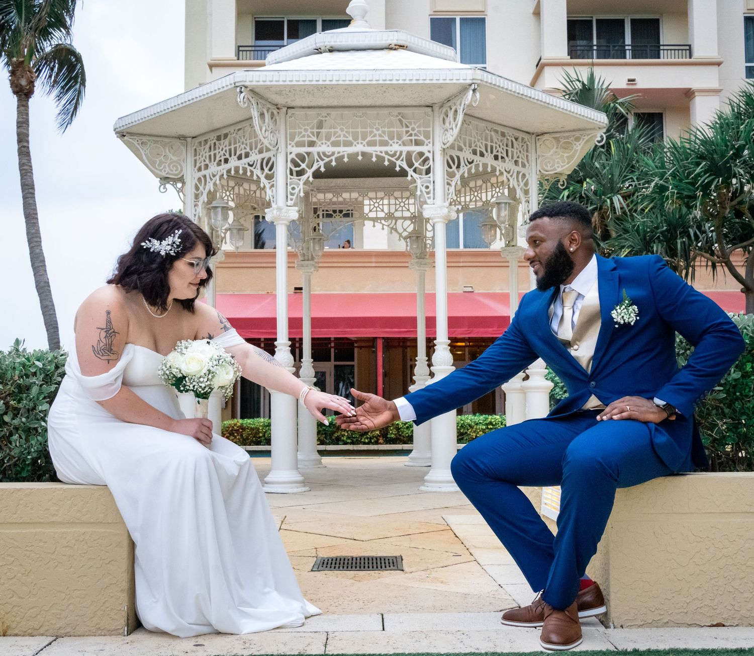 Bride and groom sit opposite sides of garden reaching for each other's hands with gazebo in background.