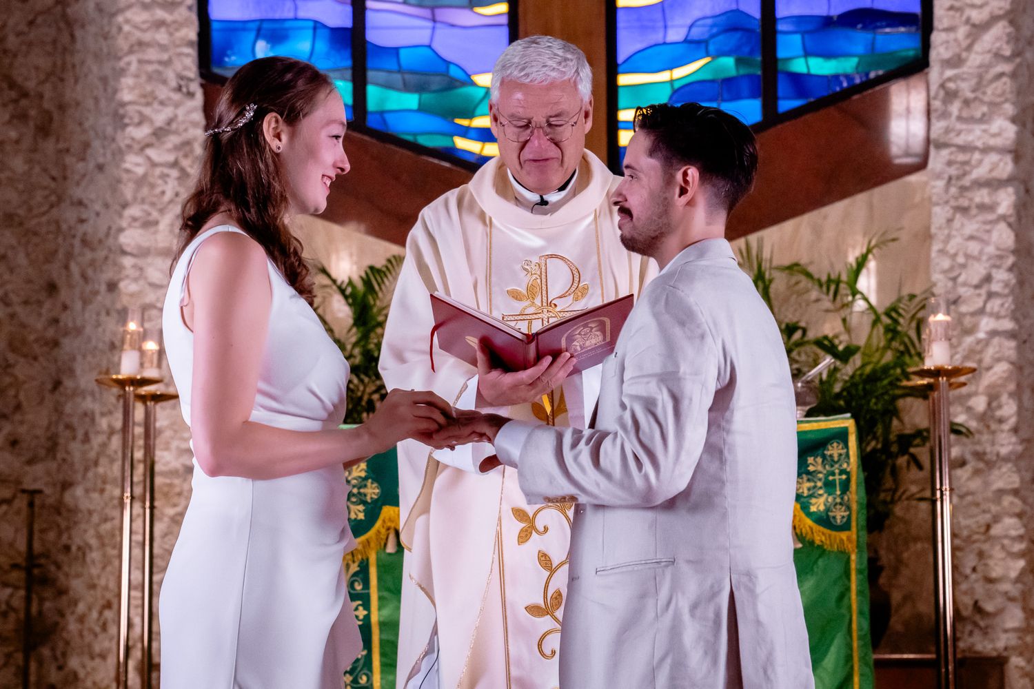 Bride and groom gaze into each other's eyes with a smile as they hold hands at alter while priest marries them.