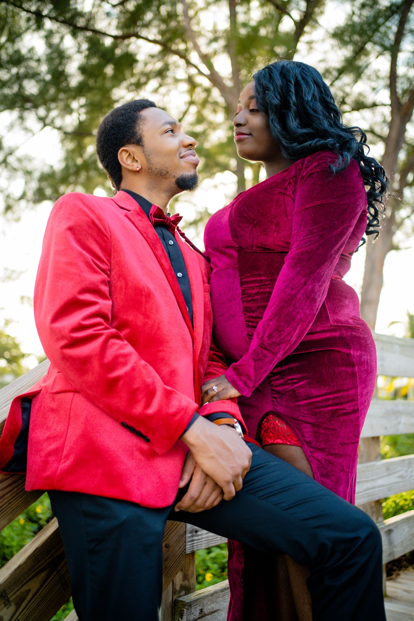 black couple in velvety suit and dress gaze into each other's eyes while on a staircase. Greenery in the background.