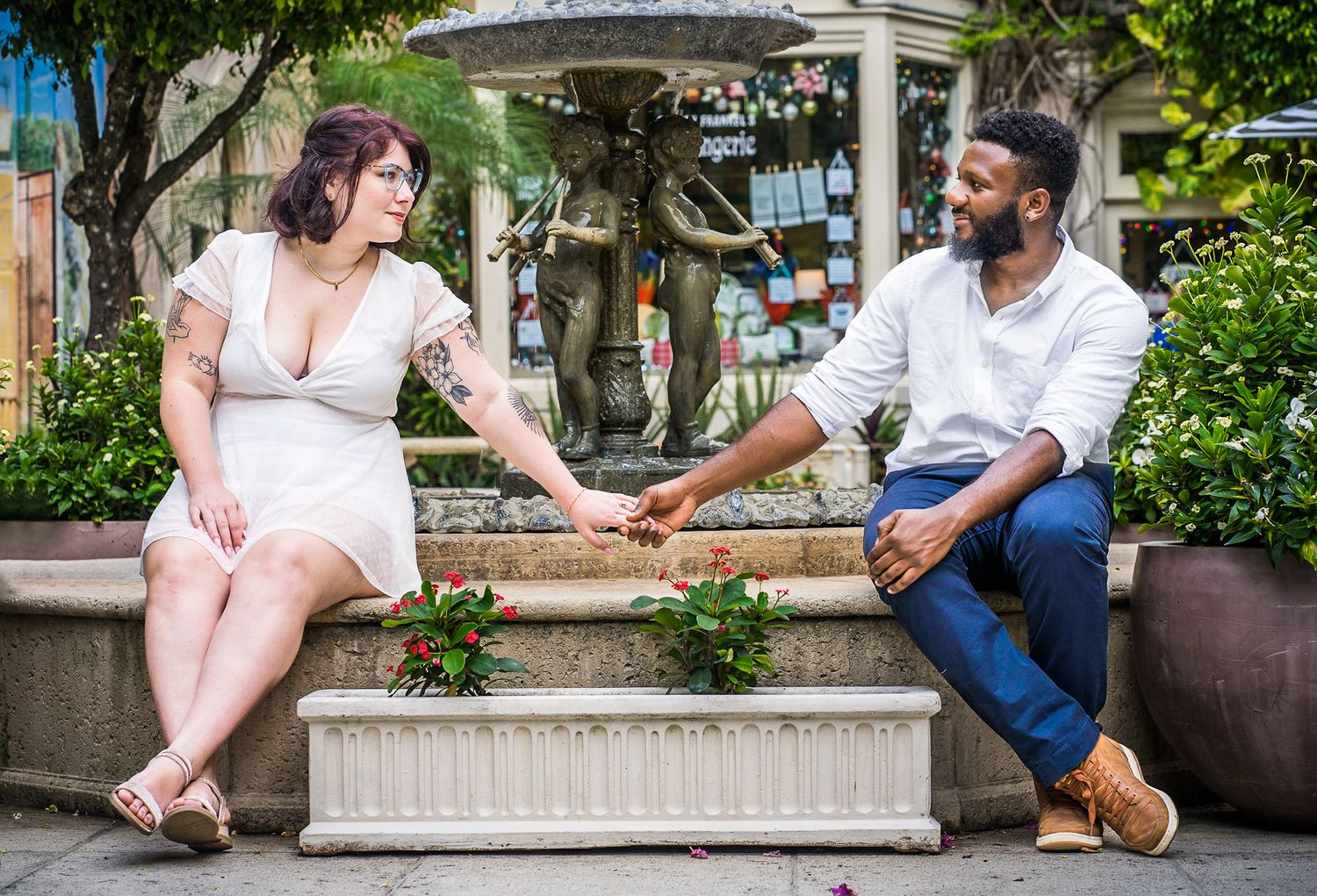 Couple holding hands while sitting at a fountain with greenery, architecture, and Mediterranean structures on palm beach island.