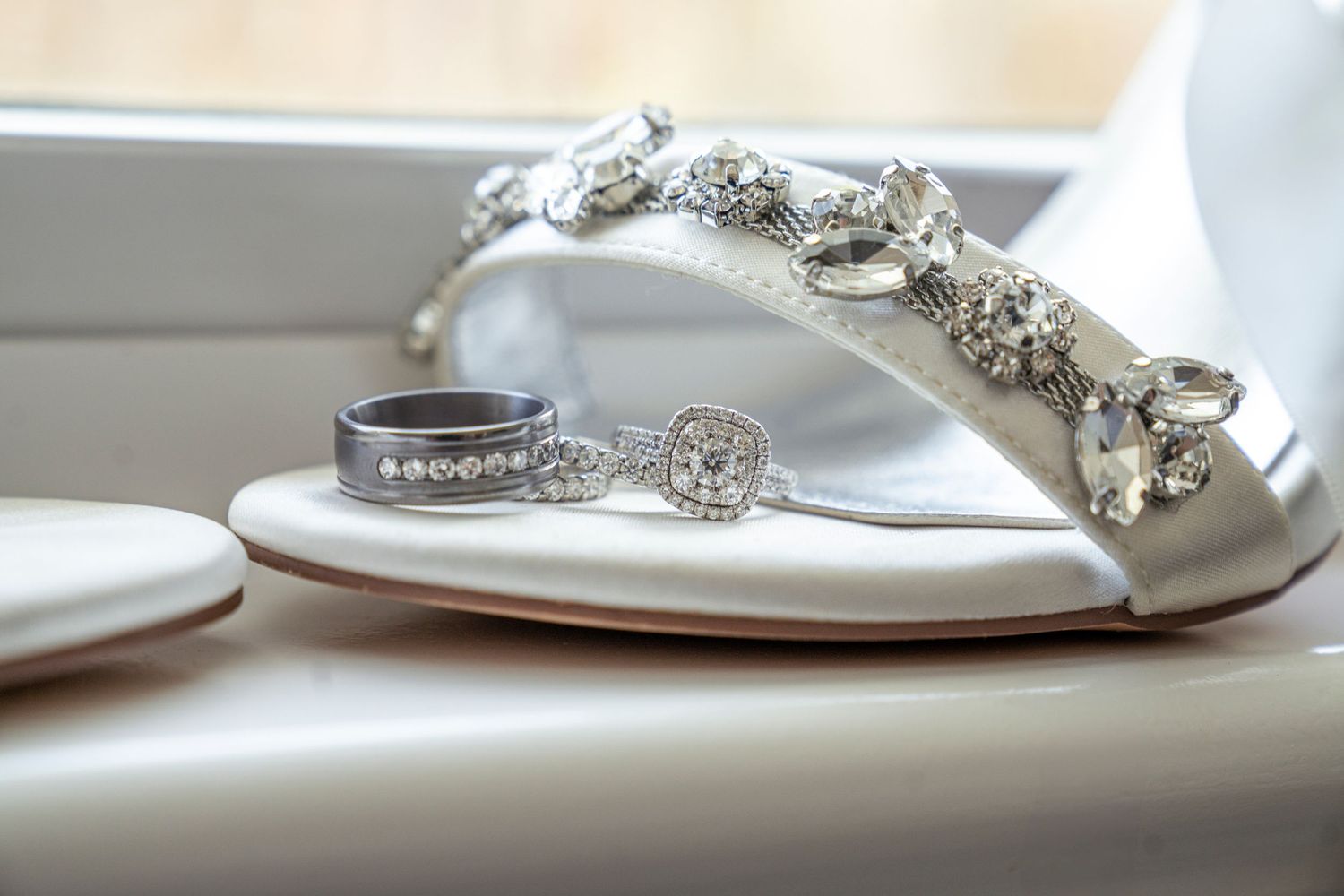 Close-up photo of bedazzled wedding shoe with both bride and groom's rings resting on a single shoe. The shoe is rested on a windowsill.