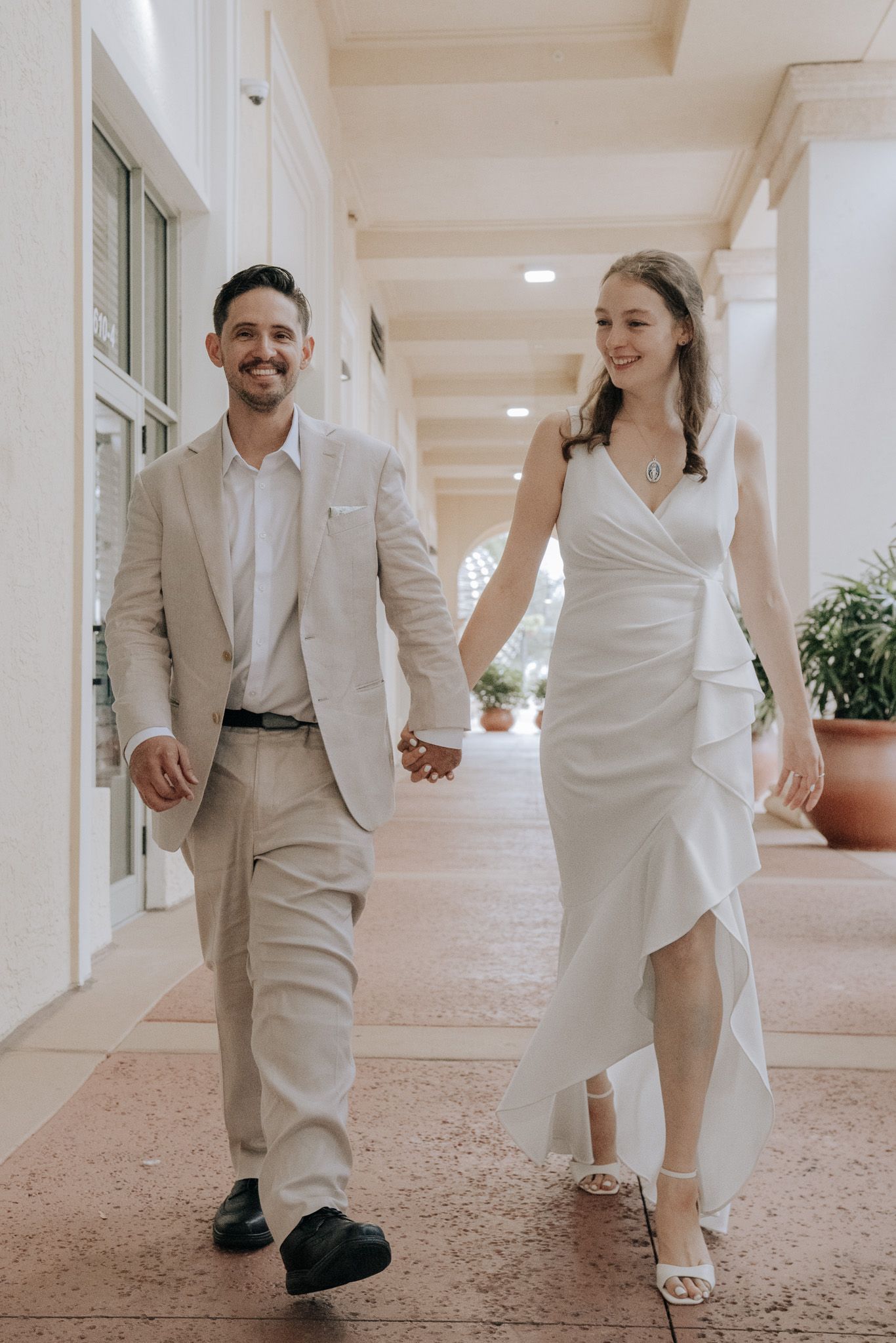 Bride and groom hold hands as they walk down an architecturally upscale area with potted greenery and white columns lining the way.
