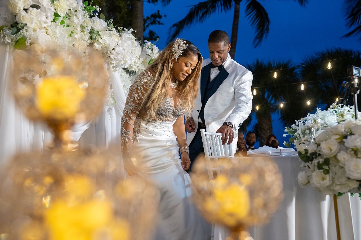 Black couple sitting down at bridal table on patio lined with white flours and blurred candles in the foreground.