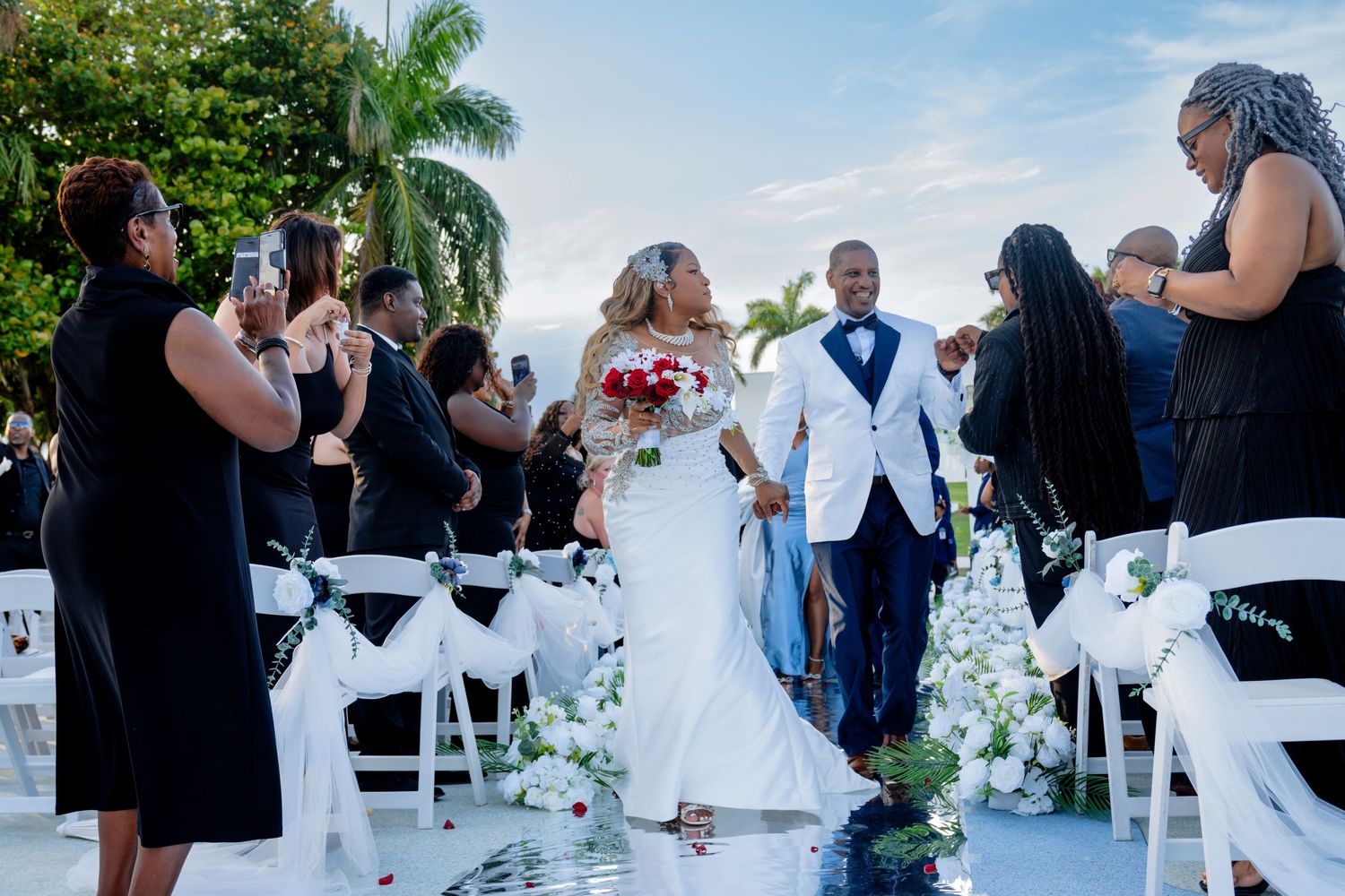 Bride and groom walk down isle, just married, as everyone stands and cheers. blue skies and trees in background.