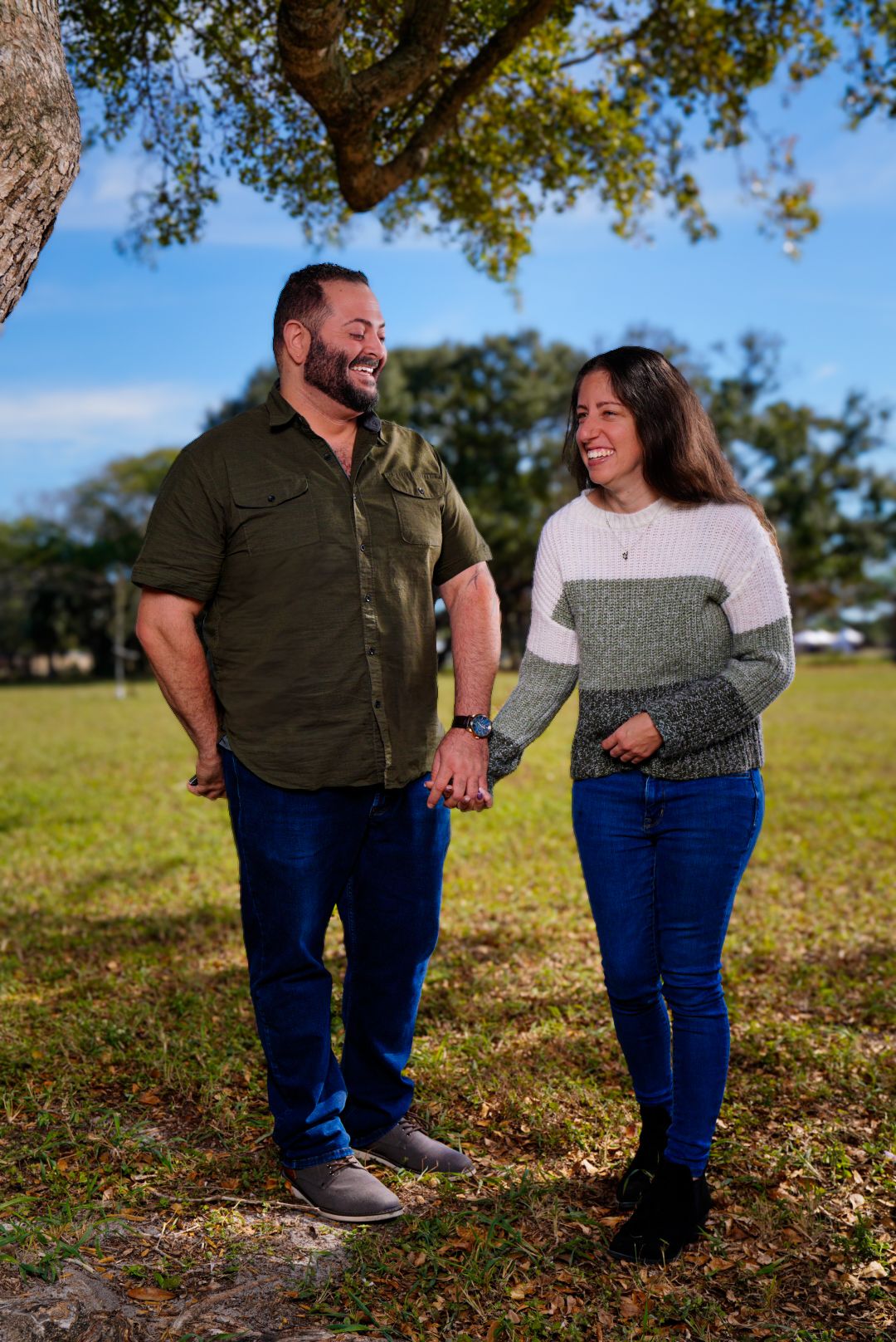 Engagement portraits of couple holding hands and laughing with tree above in foreground and blurred trees and sky in the background.