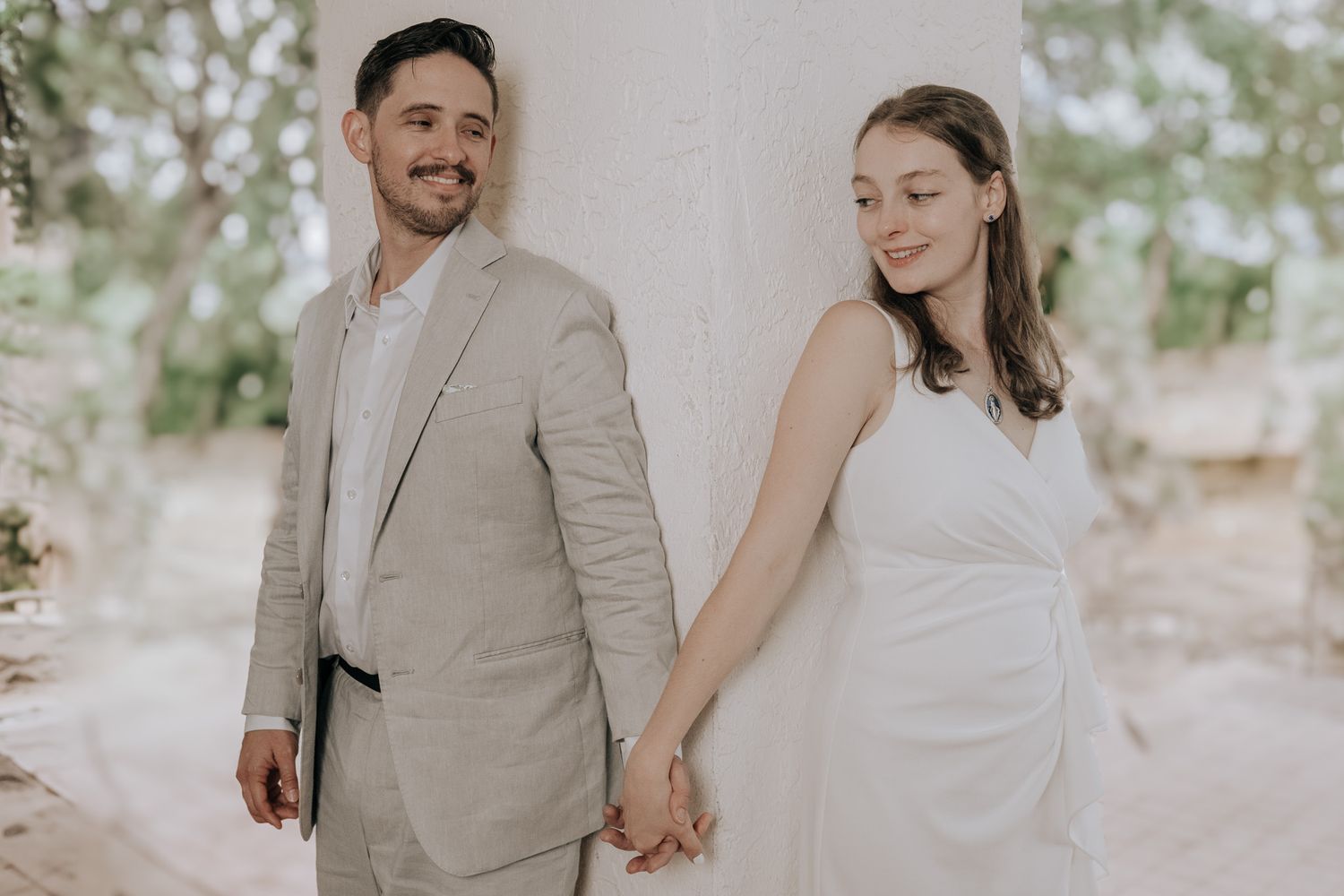 Bride and groom on each side of a column holding hands and not at each other. Behind a wall effect as they smile. Greenery in the background