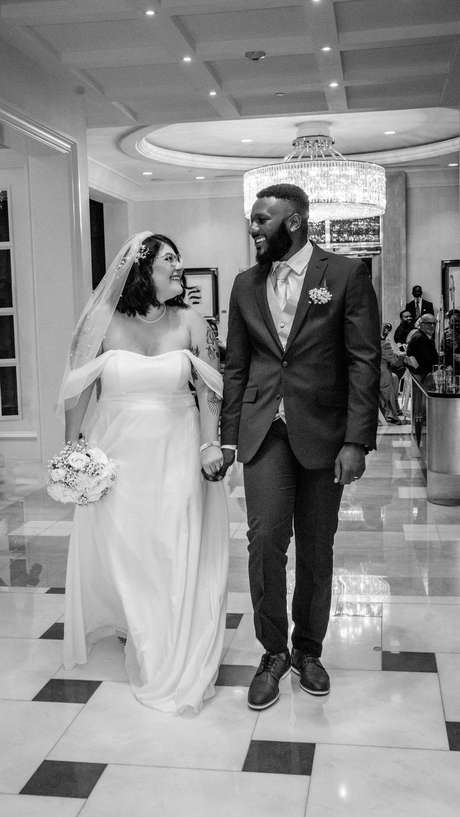 black and white photo of bride and groom smiling at each other as they walk down a corridor hand in hand.