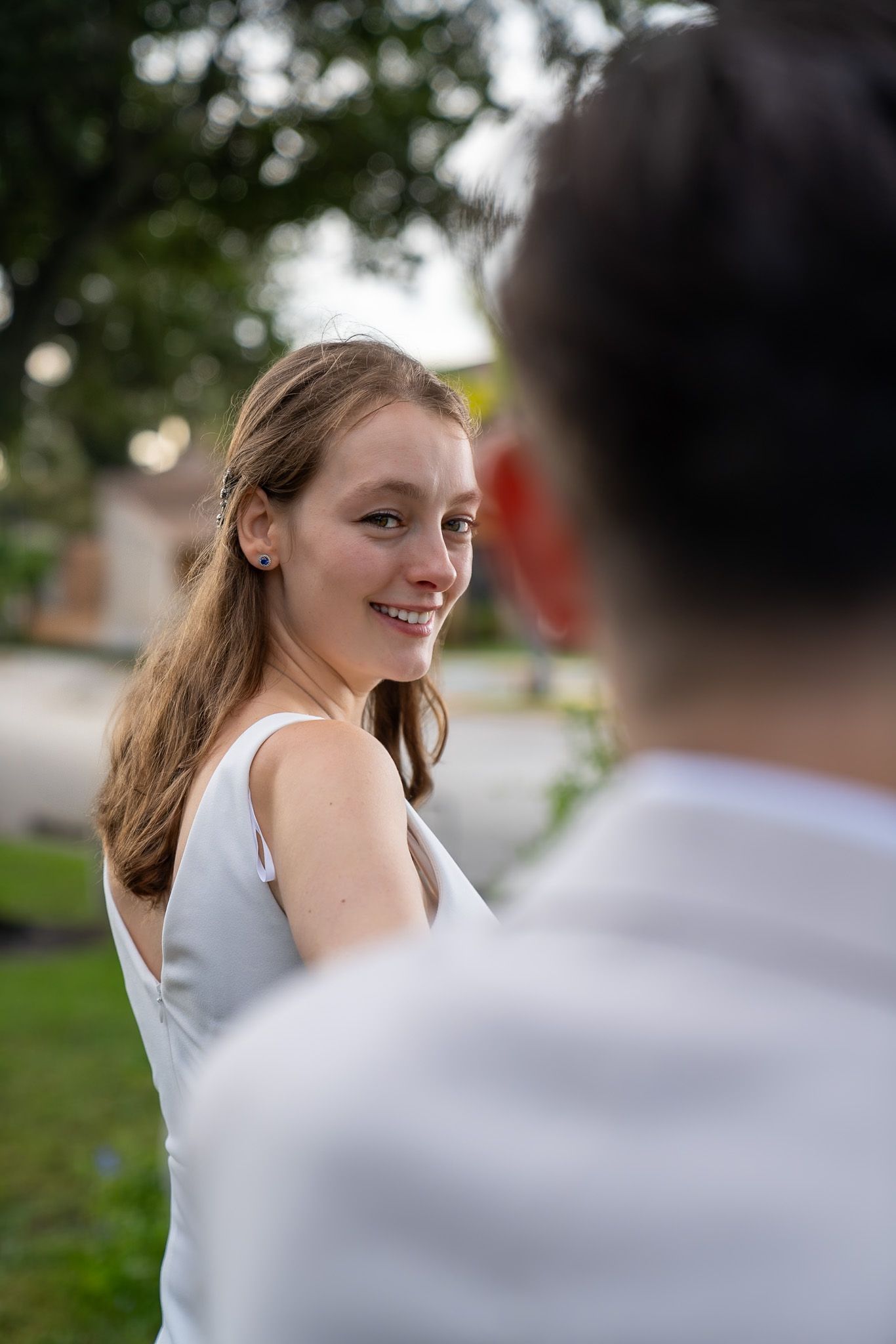 Bride gazing back towards groom with a big smile. Greenery and sky out of focus in the background.