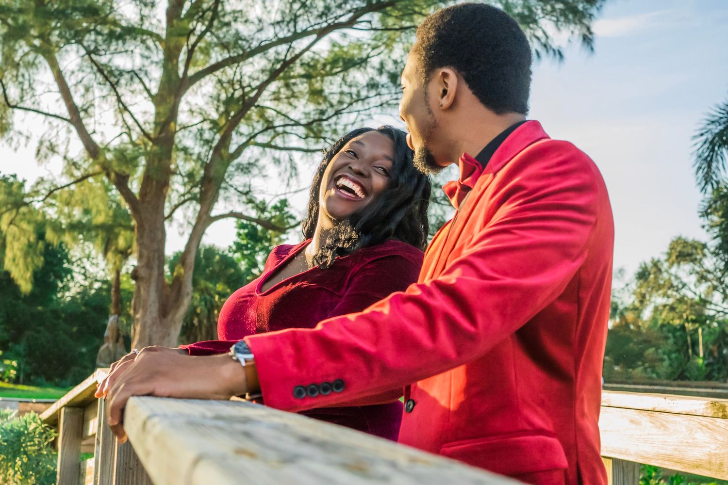 black couple in velvety jacket and dress look at each other and laugh as they rest hands on a wooden pier with trees and blue skies.