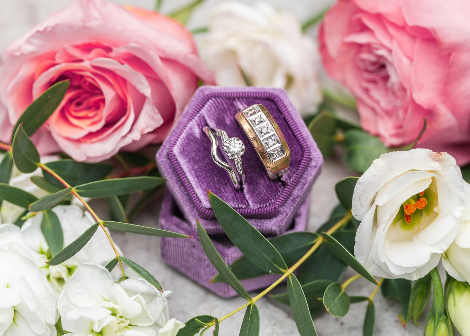 Wedding rings displayed in a purple velvet hexagon box surrounded by pink roses and white lisianthus flowers.