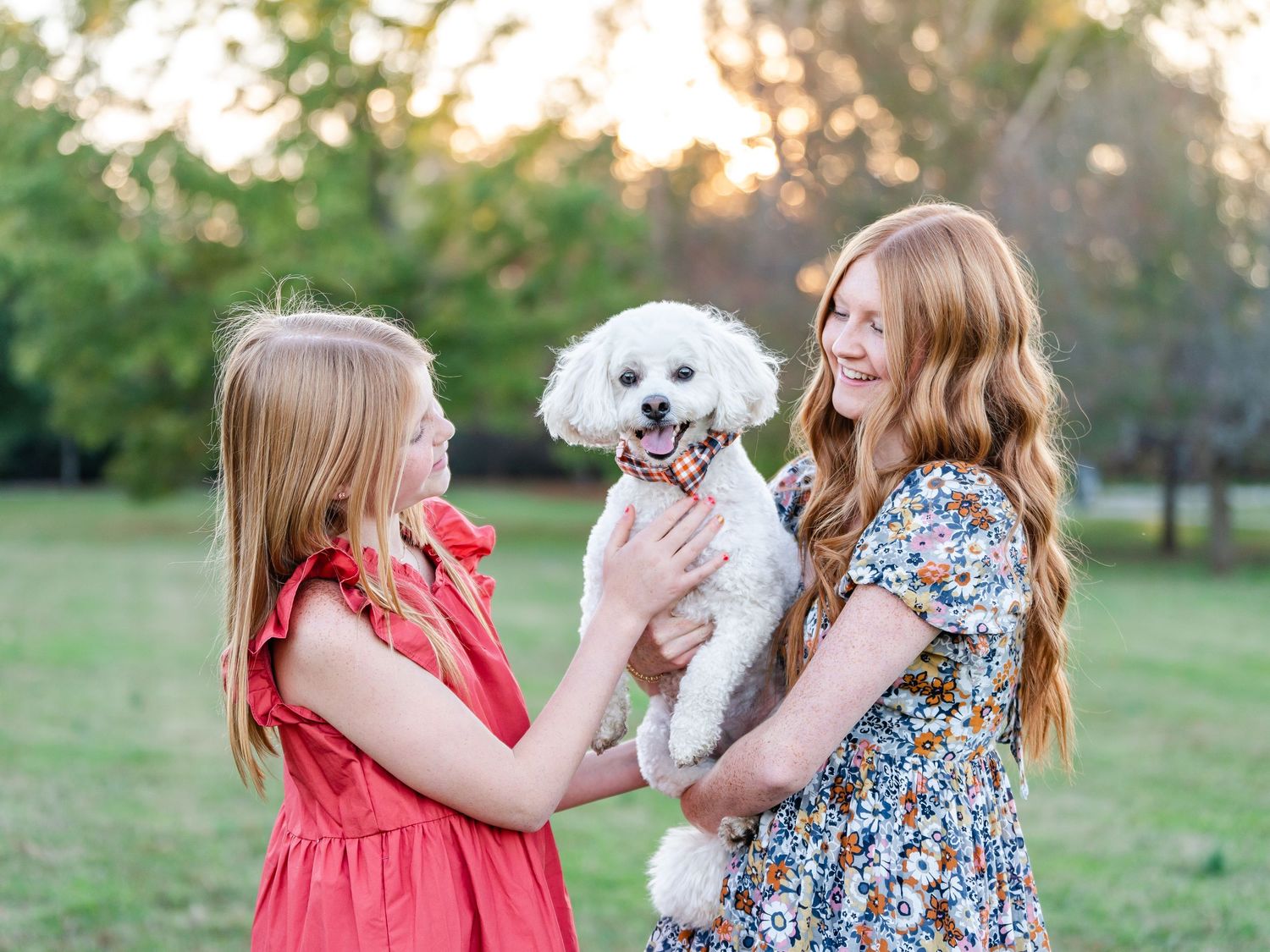 Two young girls in red and floral dresses playing with a white fluffy dog outdoors.