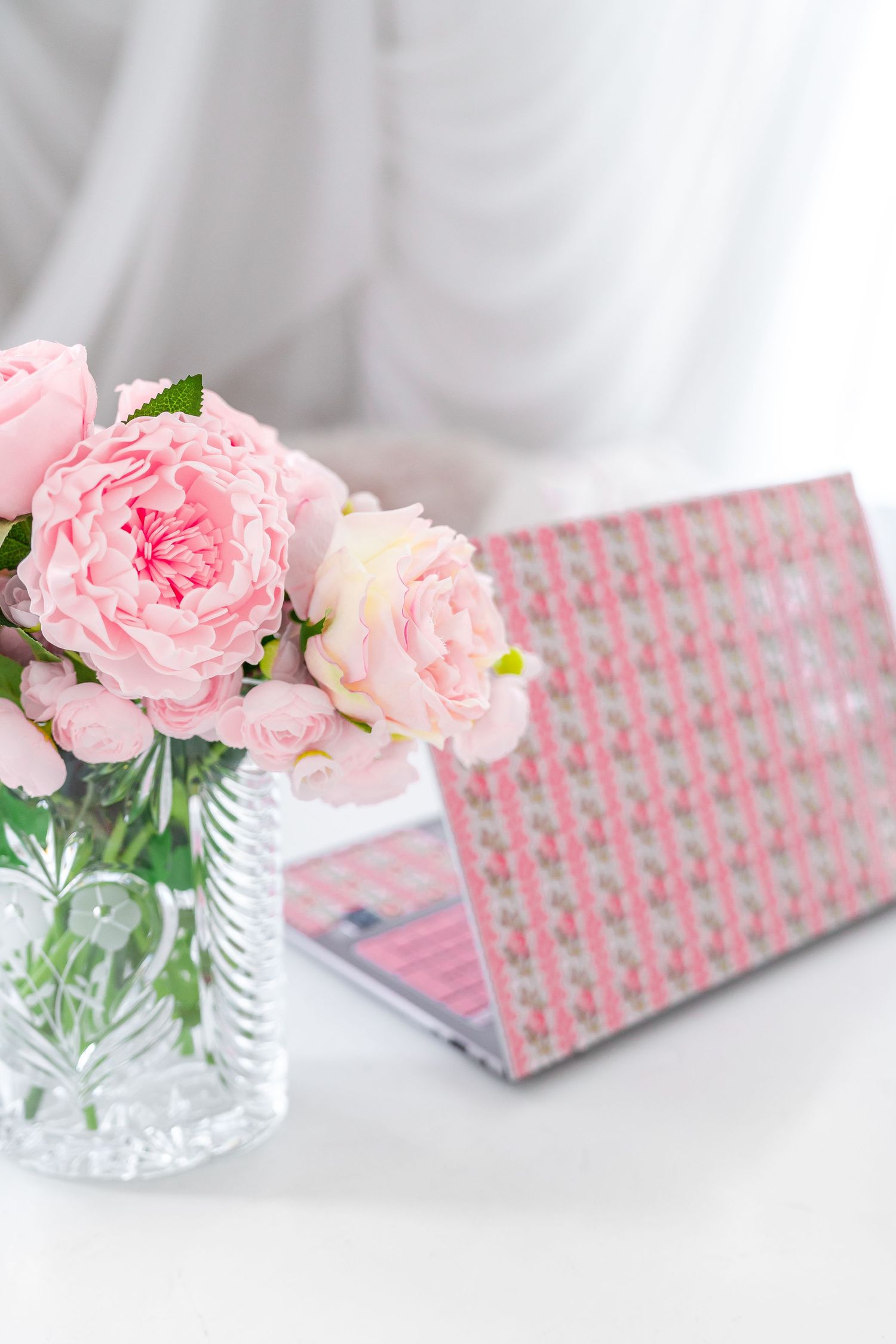 Pink roses in crystal vase beside a floral-patterned pink laptop on a white desk.