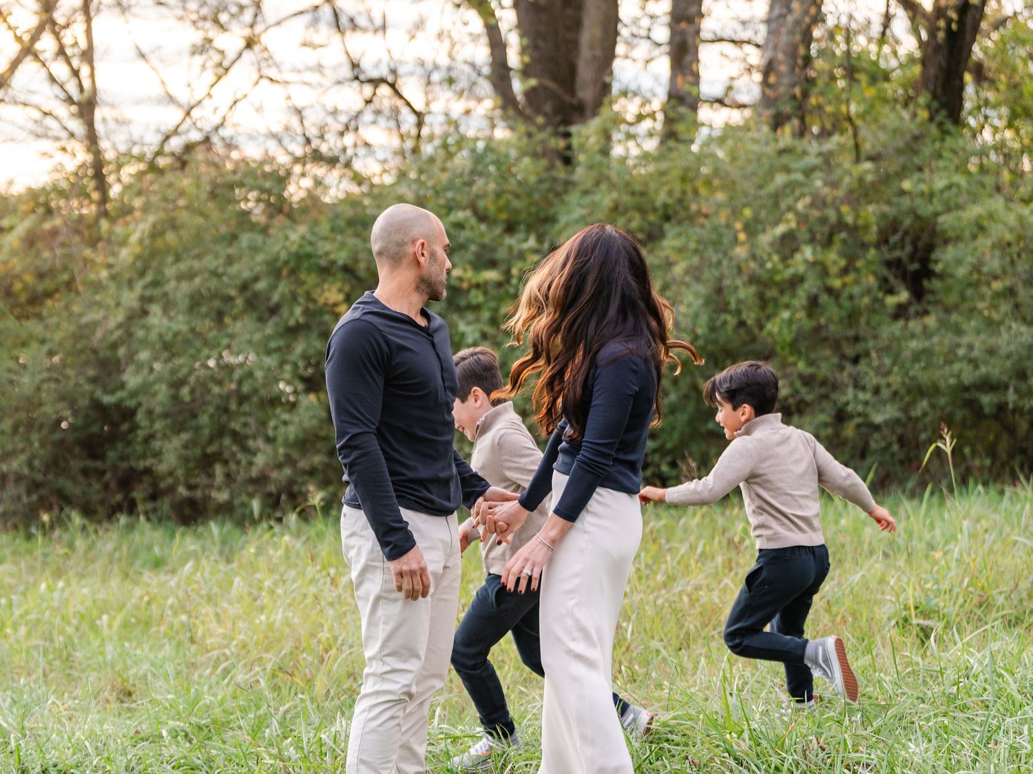 Family of four playing in a green meadow at golden hour, two children running around parents outdoors.