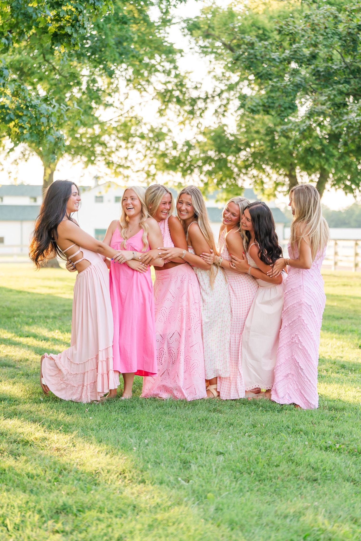 Group of six young women in pink dresses laughing and hugging together in sunny park.