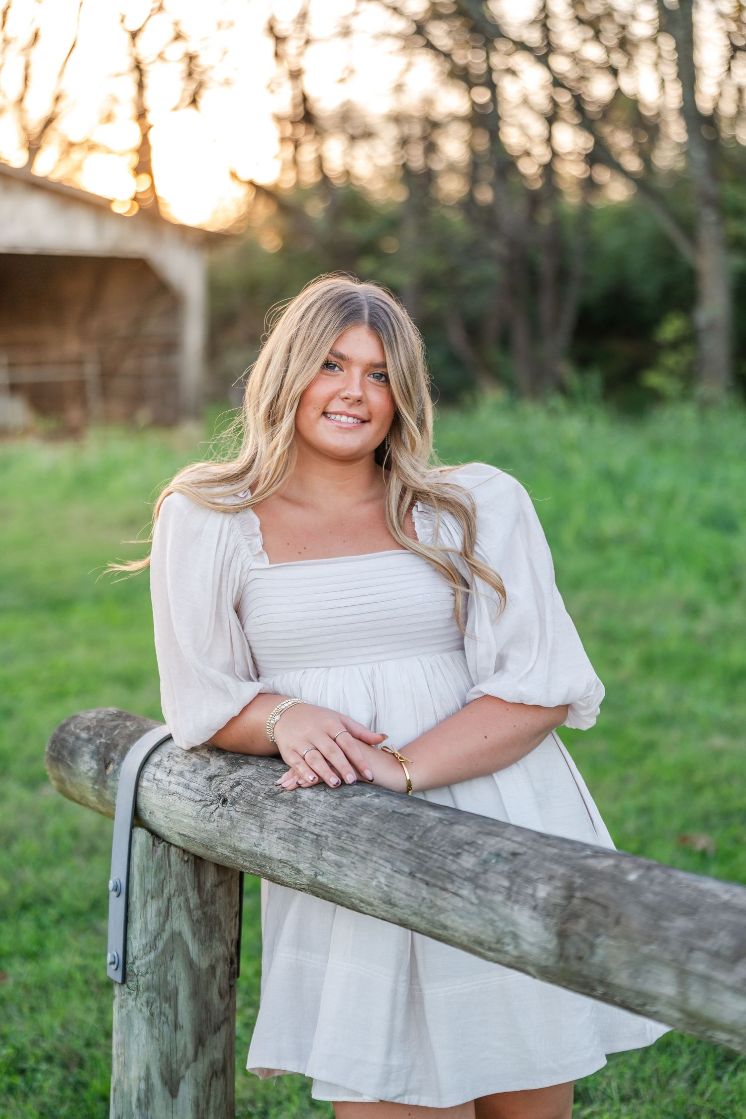 Smiling blonde woman in white dress leaning on rustic wooden fence at sunset in pastoral setting.